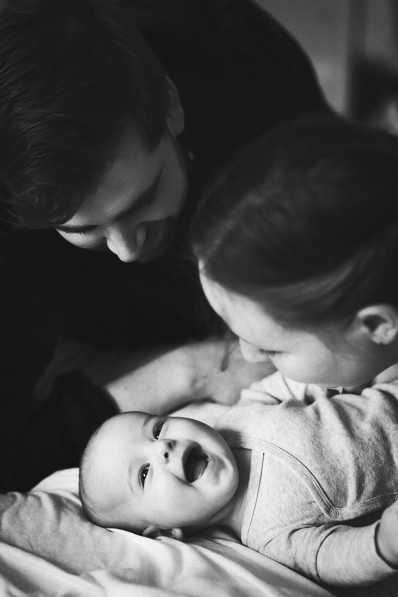 Zwei Erwachsene bücken sich zu einem lächelnden Baby, das auf einem Bett liegt.