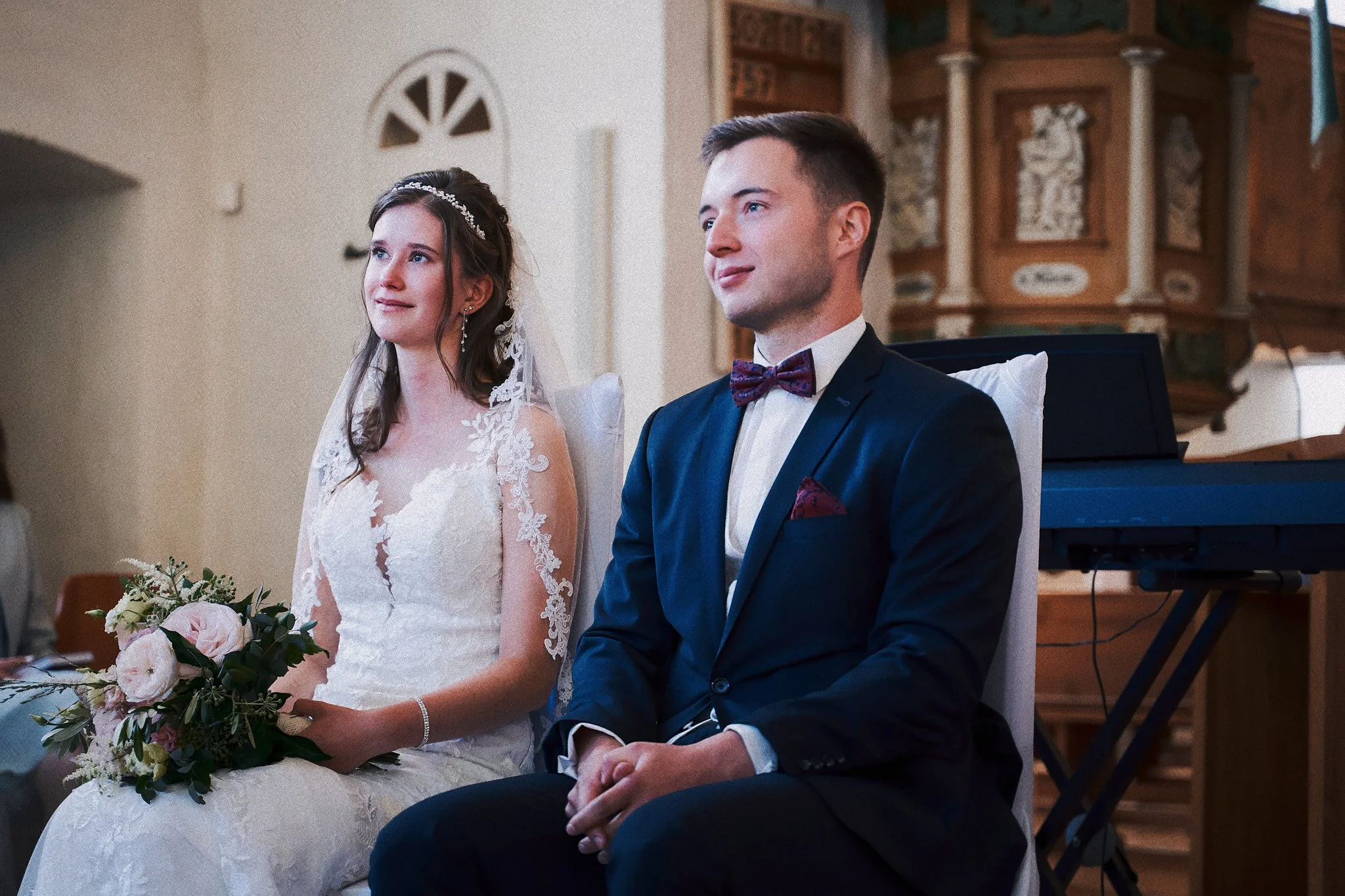 Ein Brautpaar sitzt während einer kirchlichen Zeremonie. Die Braut trägt ein weißes Spitzenkleid mit einer Tiara, hält einen Blumenstrauß, und der Bräutigam trägt einen dunklen Anzug mit Fliege.
