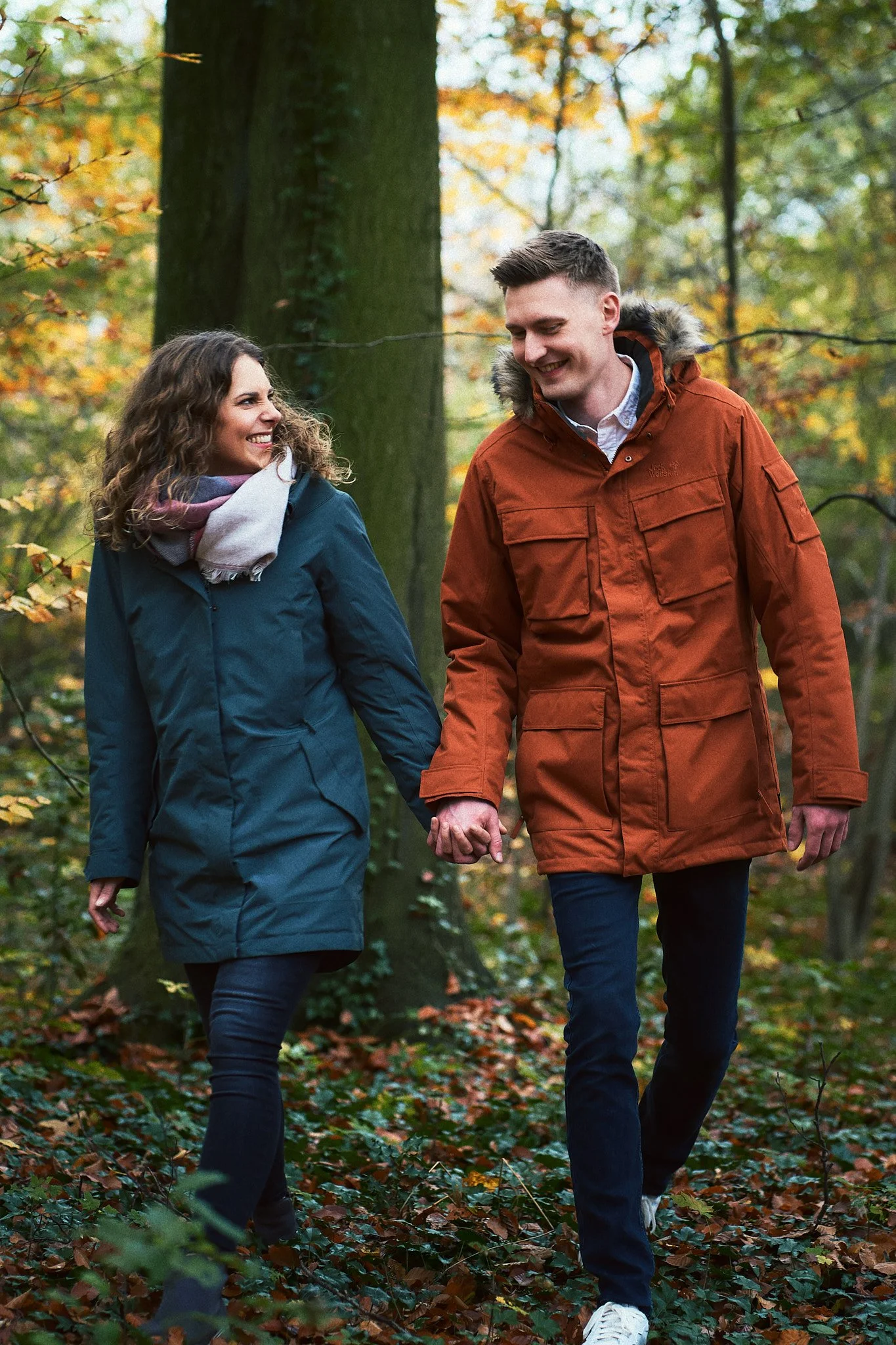 Ein Paar durchquert einen Herbstwald, Händchen haltend und lächelnd.