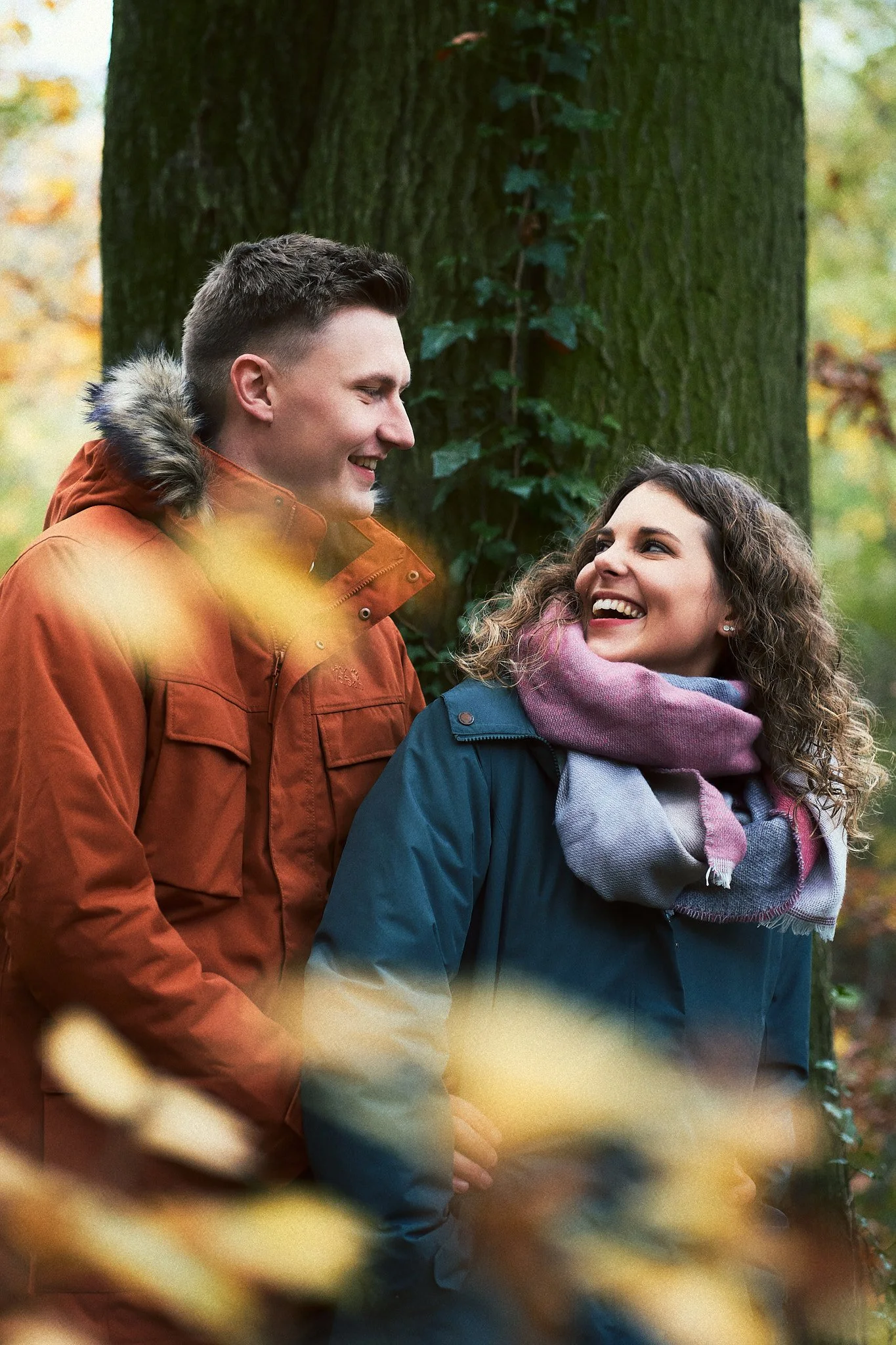 Ein Mann und eine Frau lachen und schauen sich liebevoll an, während sie im Herbstwald stehen, verkleidet mit warmen Jacken und einem Schal.