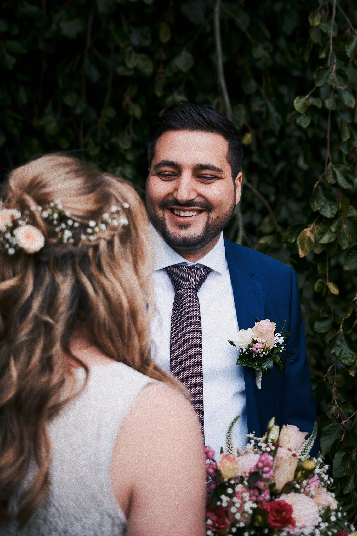 Ein Mann in einem dunkelblauen Anzug und Krawatte lächelt und schaut auf eine Frau mit lockigem, blondem Haar, die einen Brautstrauß hält, vor einem grünen Laubhintergrund bei einer Hochzeit.