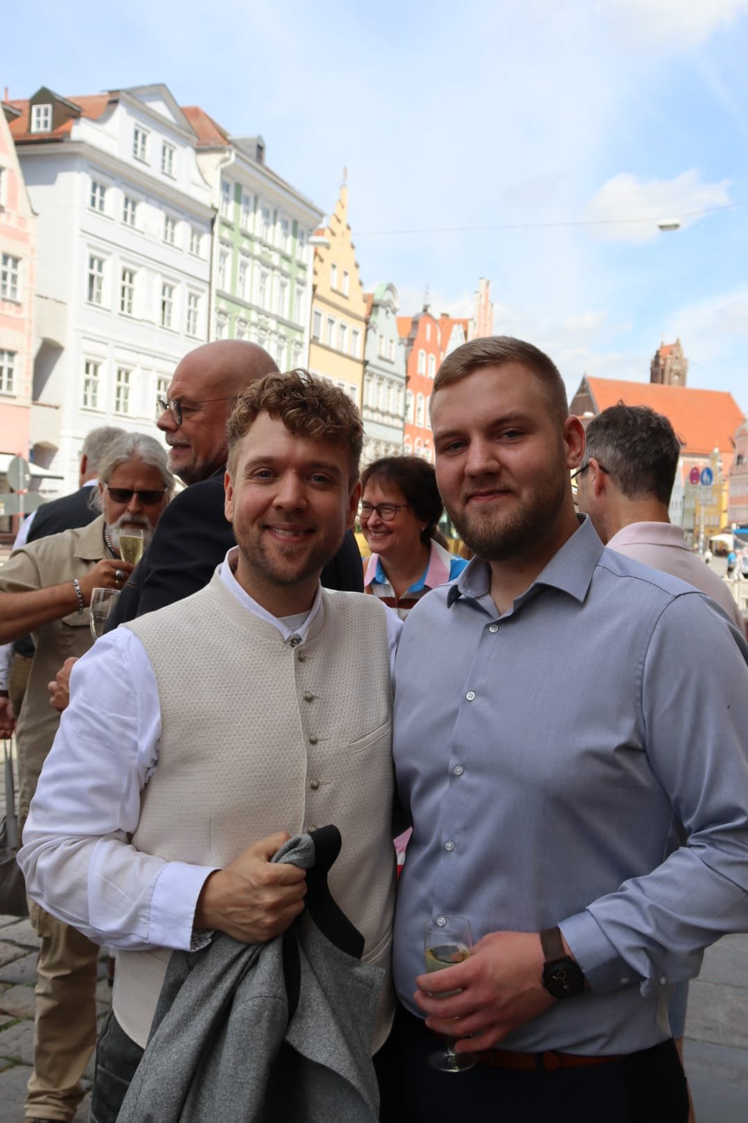 Zwei Männer posieren bei einer Feier im Freien, im Hintergrund sind weitere Personen und bunte Gebäude zu sehen.