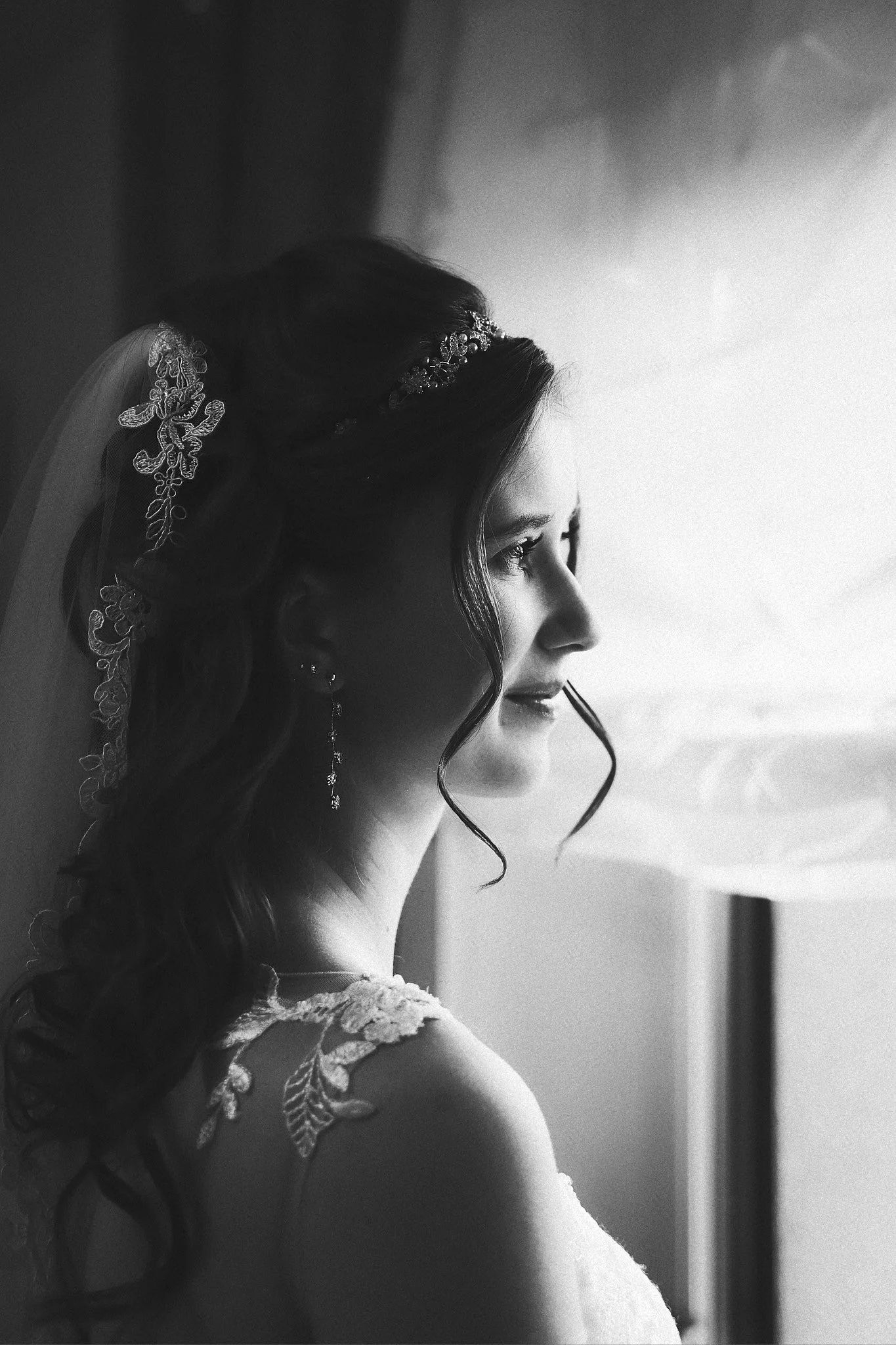 Schwarz-Weiß-Foto einer Braut, die nach links schaut, mit lockigem Haar, Haarschmuck und Ehering, vor einem Fenster.