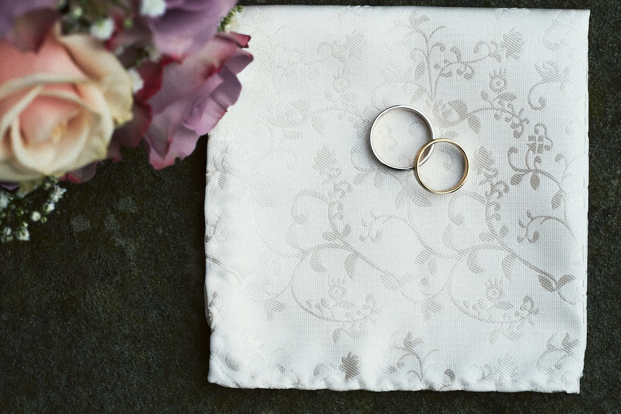 Zwei Eheringe, ein silberner und ein goldener, liegen auf einer weißen, fein gemusterten Stoffserviette. Im linken Bildrand ist ein Paar Blumen in Pastellfarben sichtbar, hervorgehoben sind dunkelrosafarbene Rosen und kleine weiße Blumen.