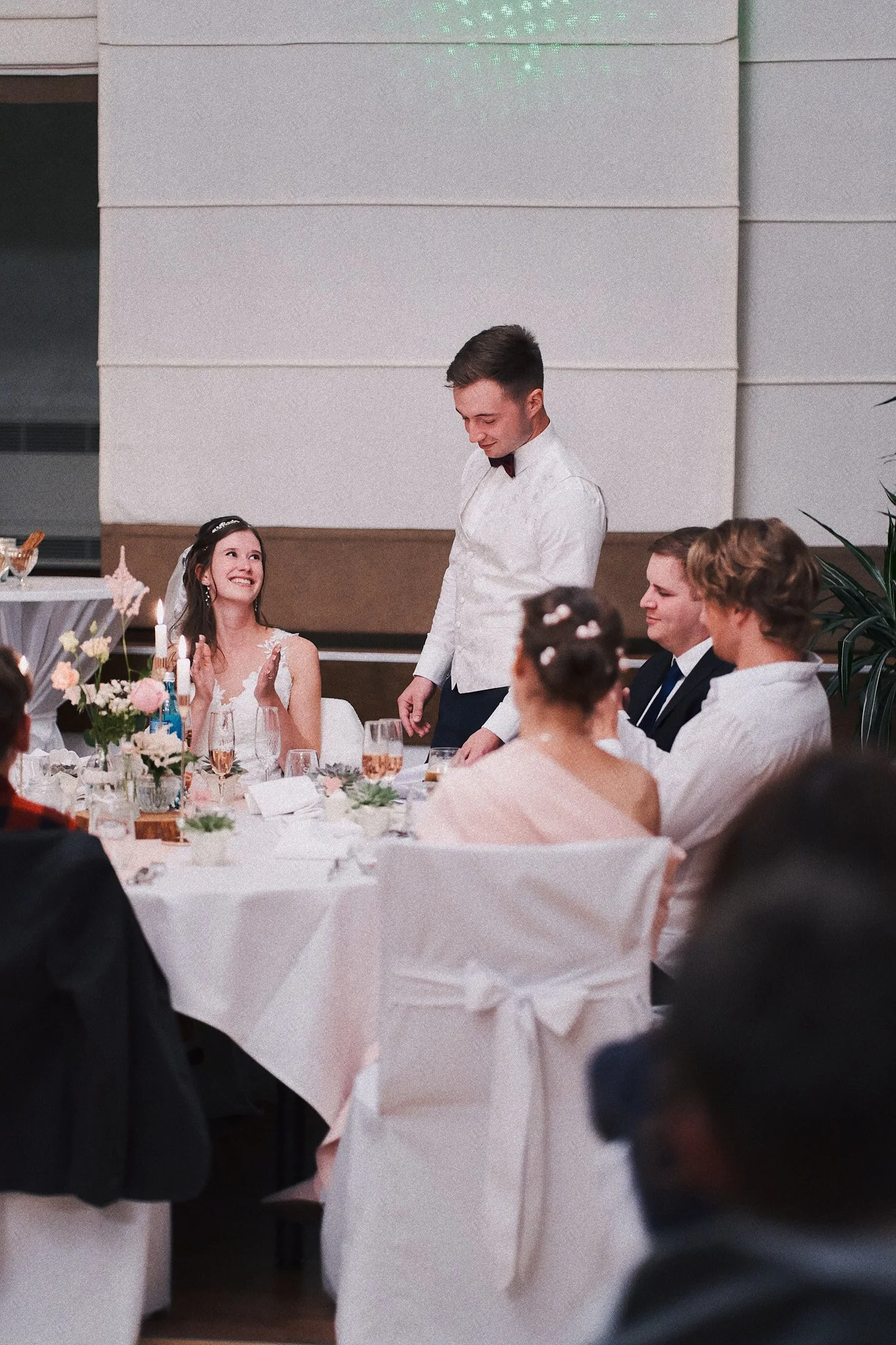Ein Hochzeitsessen mit mehreren Gästen, einem Brautpaar und einer Dekoration mit Blumen.
