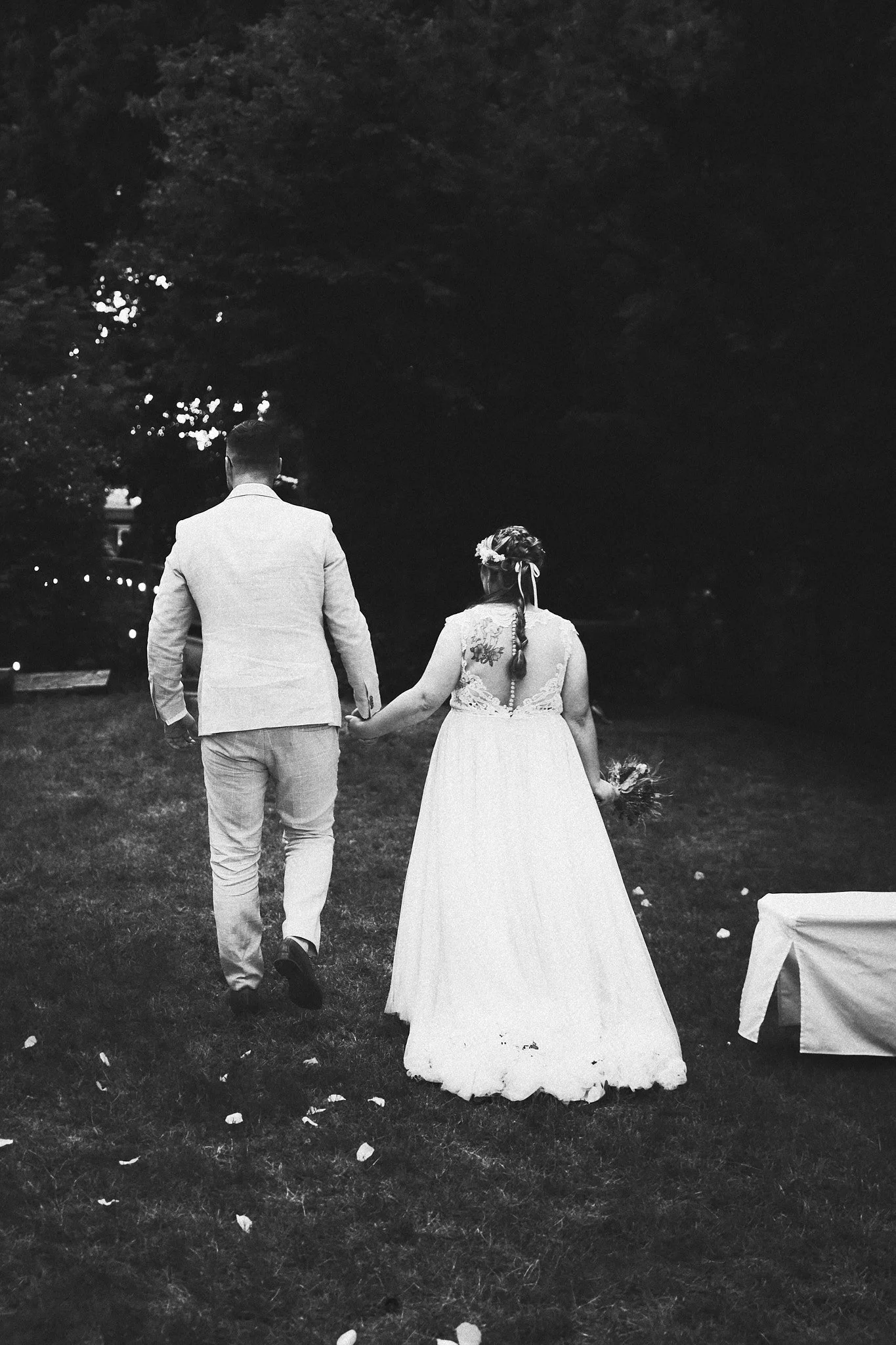 Ein Brautpaar, das Hand in Hand auf einer Wiese bei Nacht geht, mit Bäumen im Hintergrund. Die Frau trägt ein langes weißes Brautkleid und einen Blumenkranz, der Mann trägt einen Anzug. Es ist eine schwarz-weiße Fotografie.
