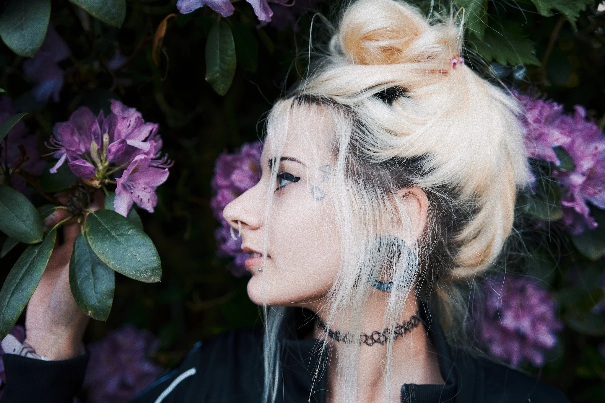 Eine junge Frau mit blonden, gefärbten Haaren und Tattoos am Gesicht, die an einem Busch mit lila Blumen riecht.