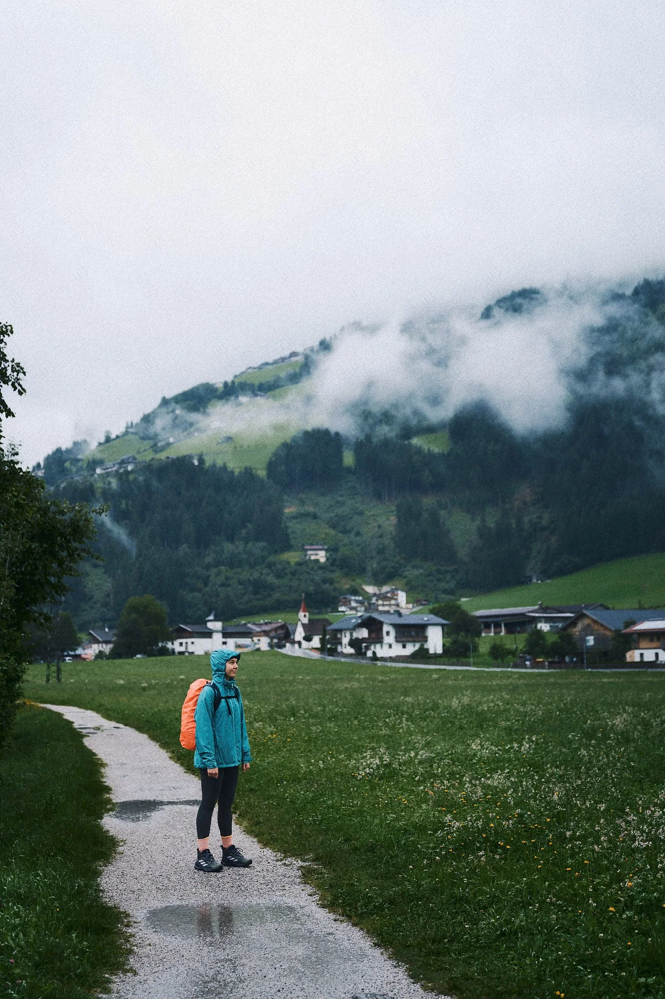 Eine Person mit Rucksack steht auf einem schmalen Weg in einer grünen Landschaft, im Hintergrund ein Dorf und nebelverhüllte Berge.