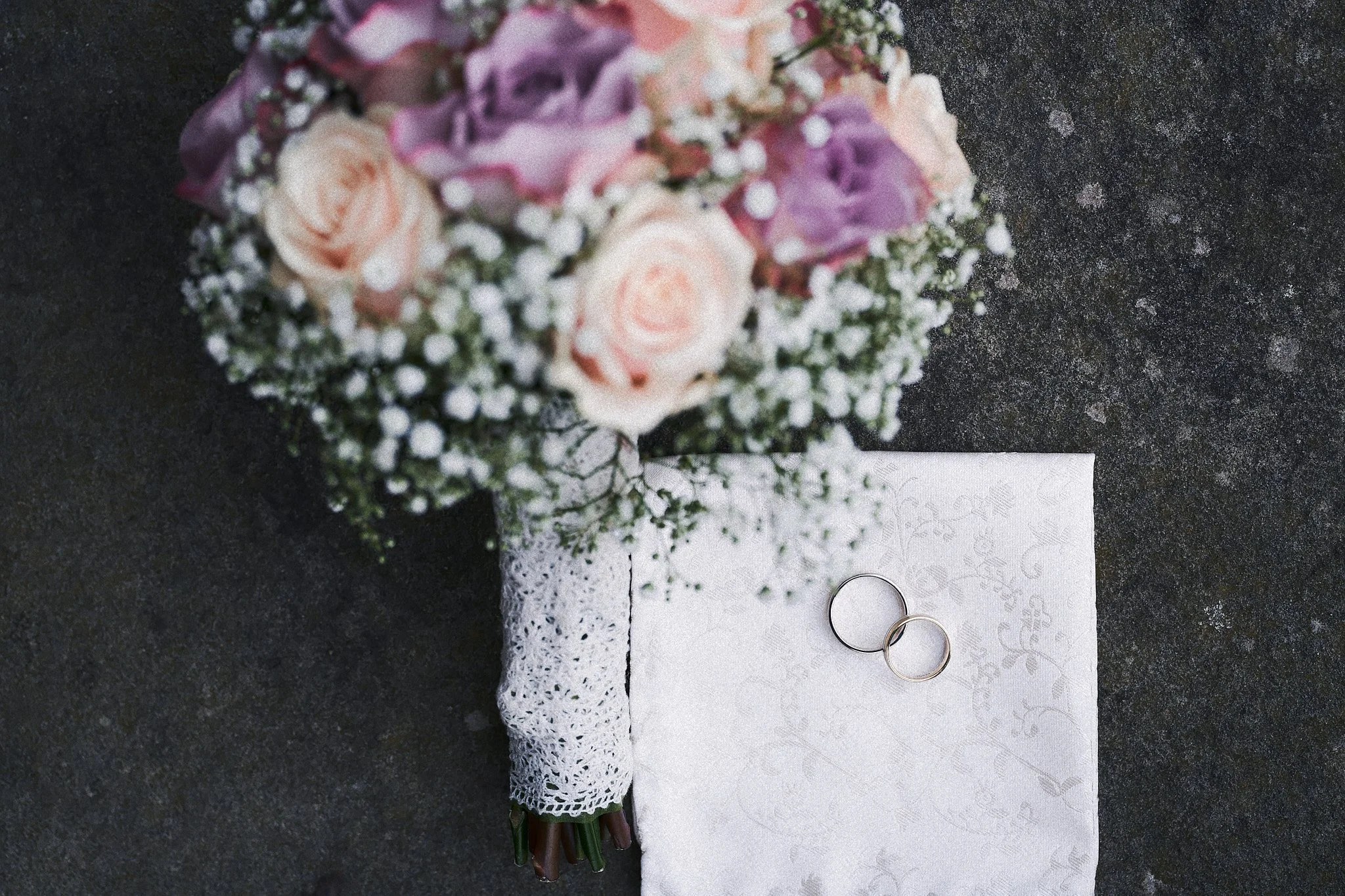Ein Blumenstrauß aus rosa und lilafarbenen Blumen, begleitet von weißen Baby’s Breath, liegt auf einer schwarzen Oberfläche neben Hochzeitsringen auf einer weißen Tischdecke.