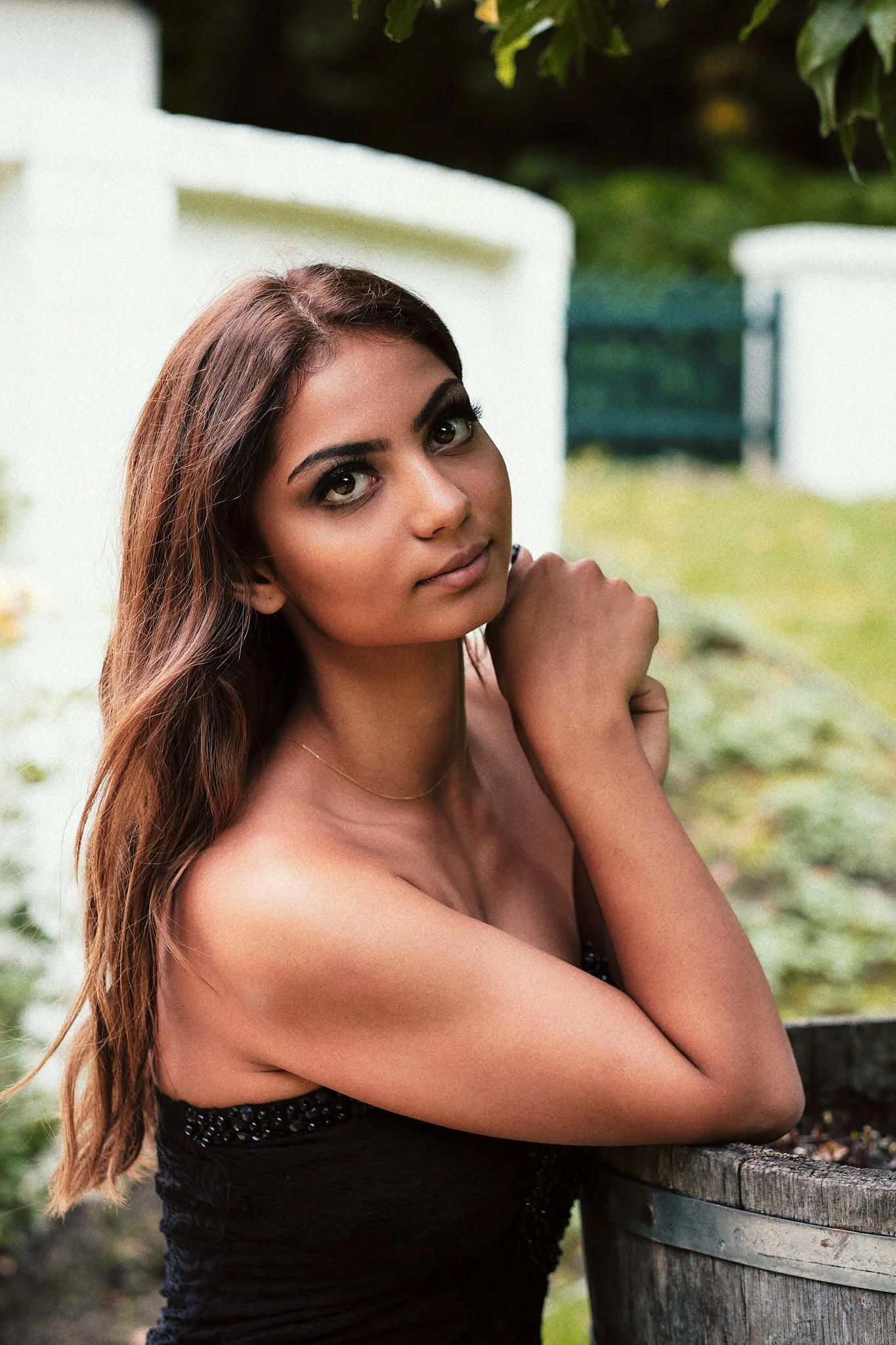 Eine junge Frau mit langen braunen Haaren und dunklem Make-up sitzt an einem runden Holztisch im Freien, umgeben von grünen Pflanzen.