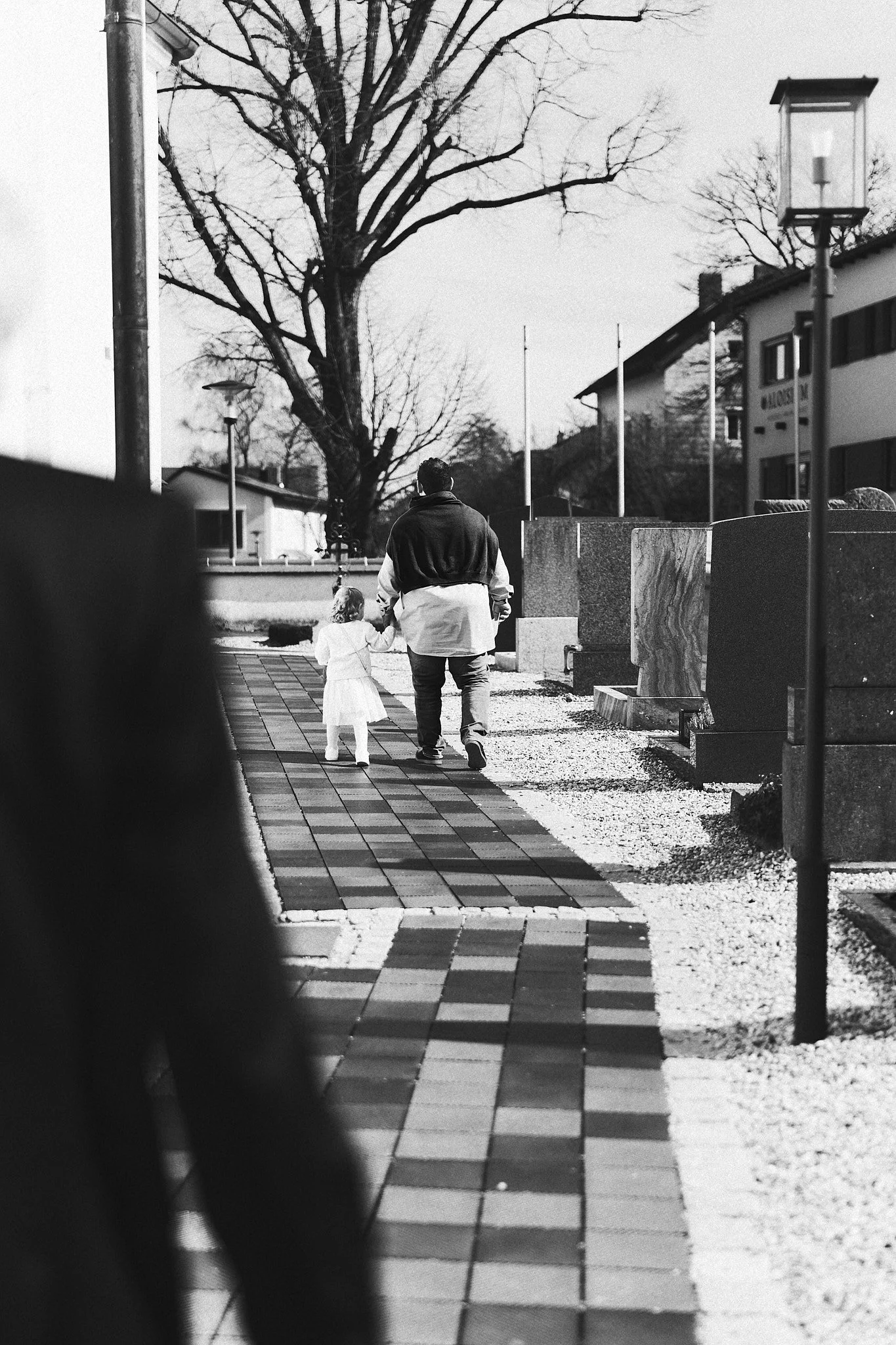 Zwei Menschen, ein Erwachsener und ein Kind, gehen Hand in Hand auf einem gepflasterten Weg im Freien, mit Bäumen und Gebäuden im Hintergrund, in schwarz-weißer Fotografie.