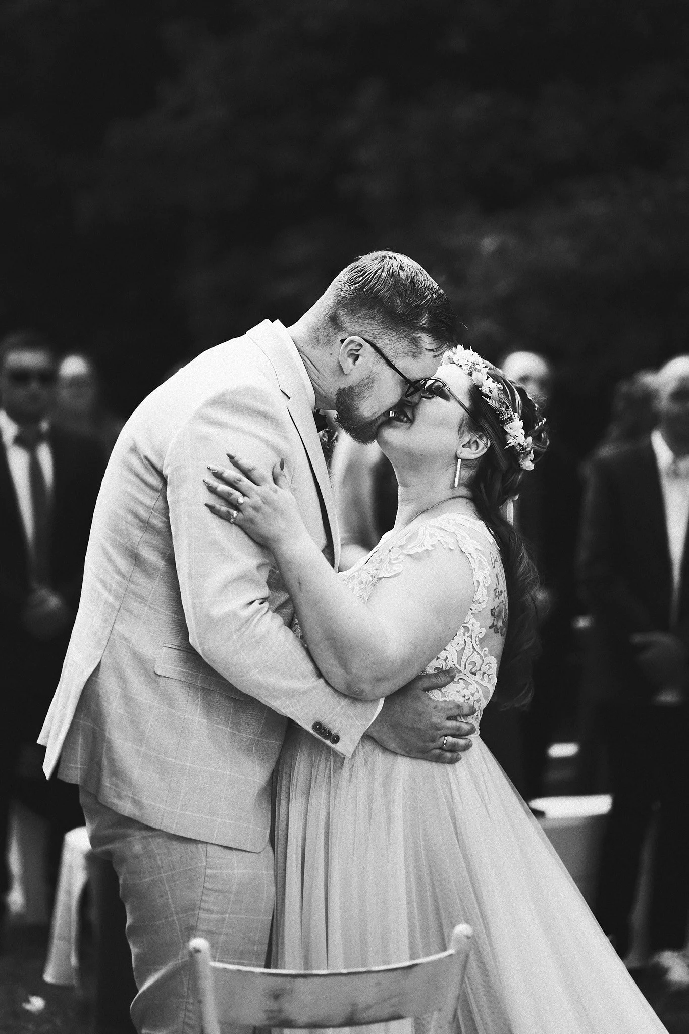 Ein Brautpaar bei der Hochzeit, umarmt sich und küsst sich leidenschaftlich, im Hintergrund sitzen Gäste, schwarz-weiß Fotografie.