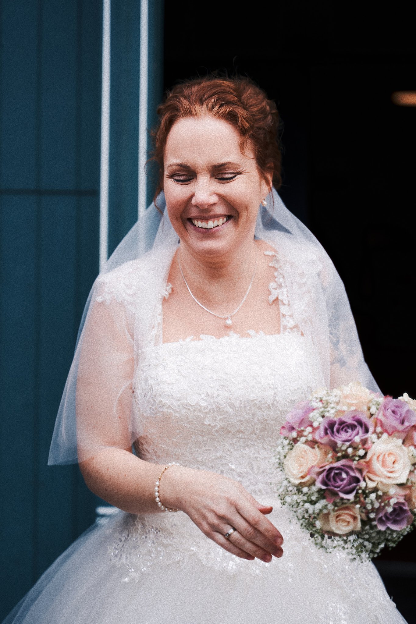 Eine lächelnde Braut in einem weißen Hochzeitskleid mit Perlenschmuck und Brautstrauß in den Händen.