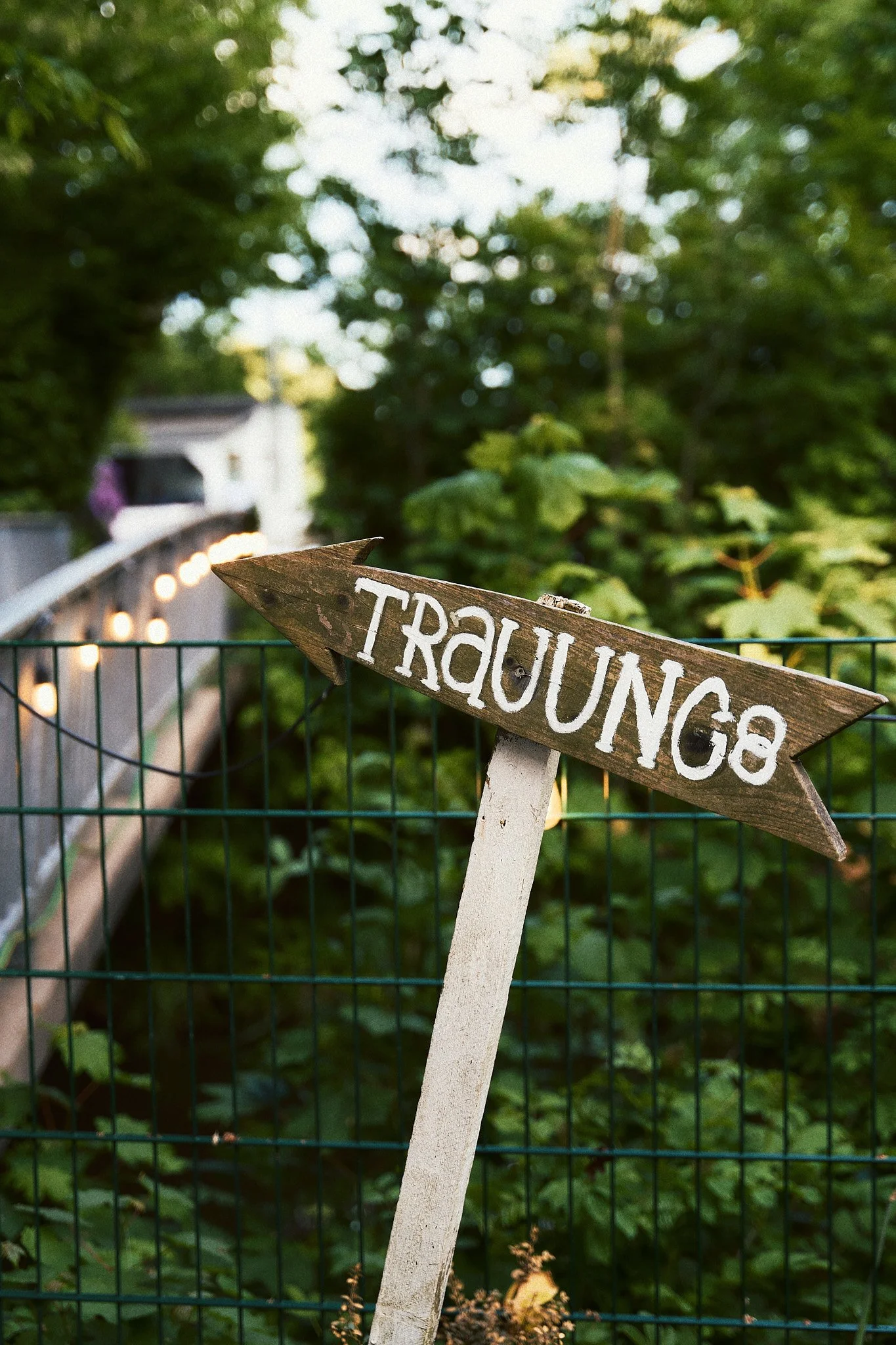 Ein handgeschnitztes Holzschild mit der Aufschrift 'Trauung' zeigt nach links, im Hintergrund grünes Laub und eine Gartenumzäunung.