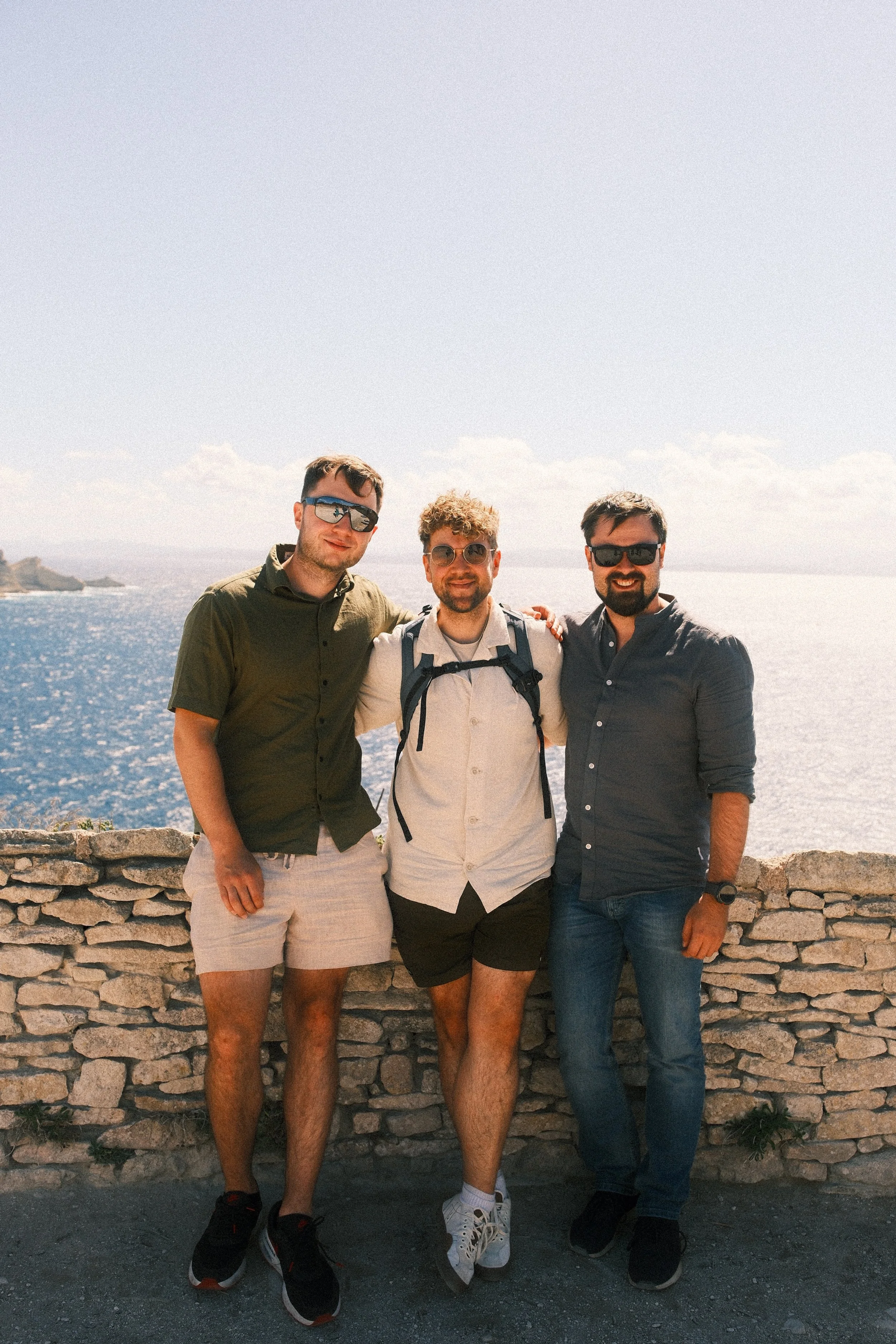 Drei Männer stehen an einer Mauer am Meer und lächeln, während sie die Sonne genießen.