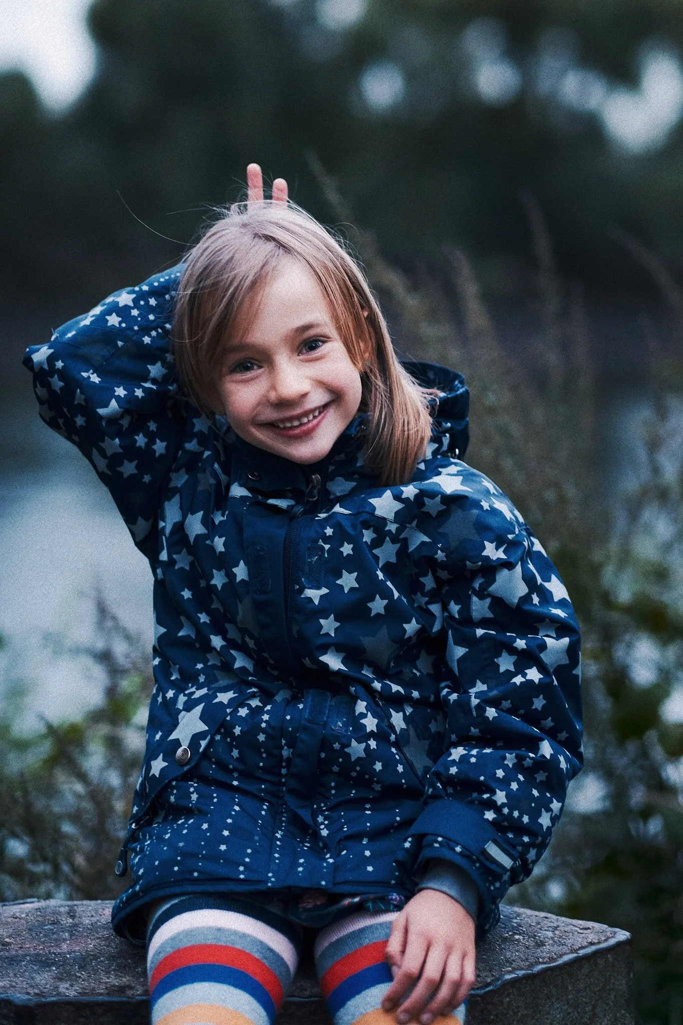 Ein lachendes Mädchen mit roten Haaren in einer blauen Jacke mit Sternmuster, sitzt auf einem Stein im Freien.