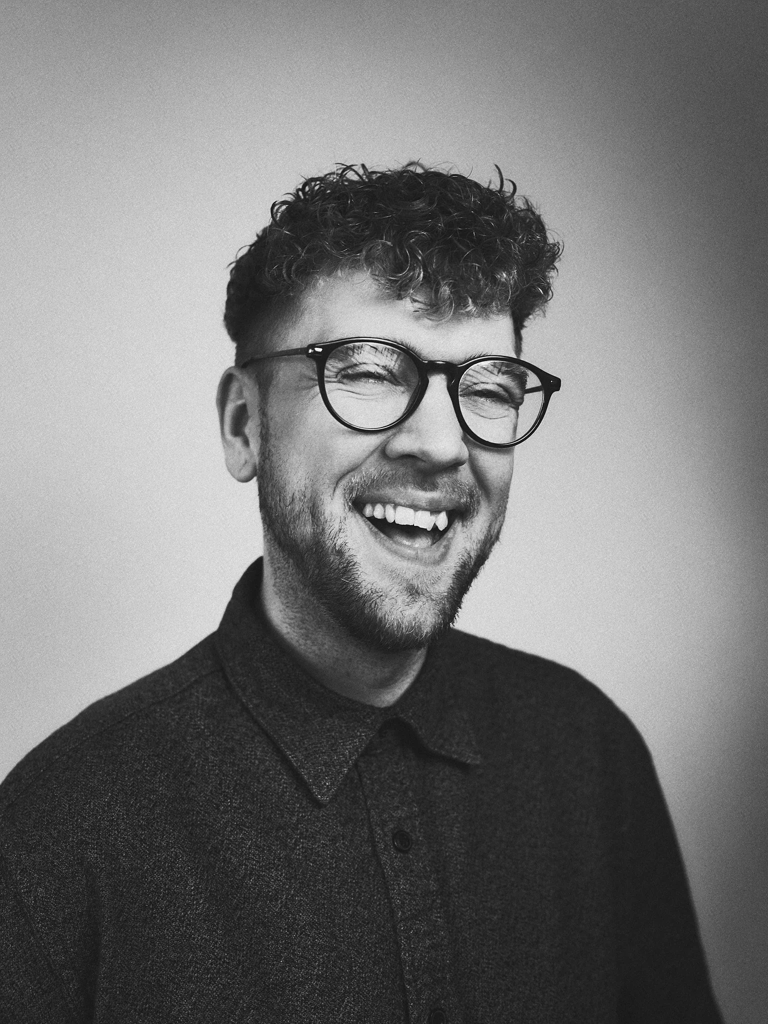 Schwarz-Weiß-Foto eines lachenden Mannes mit Brille und kurzen, lockigen Haaren, trägt einen dunklen Pullover mit Knopfleiste.