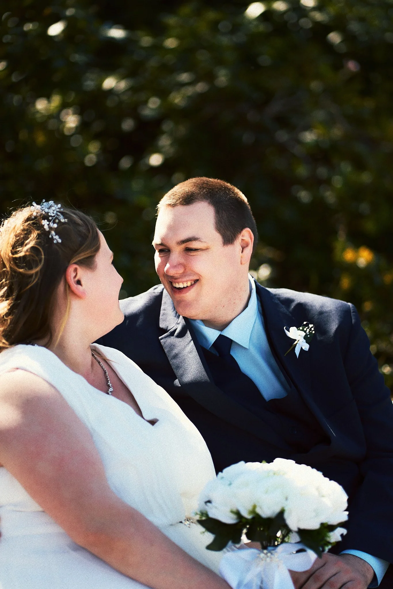 Aufnahme eines glücklichen Paares bei ihrer Hochzeit im Freien, die Braut trägt ein Weißes Kleid mit Blumen im Haar, der Bräutigam trägt einen schwarzen Anzug mit Krawatte, beide schauen sich liebevoll an.