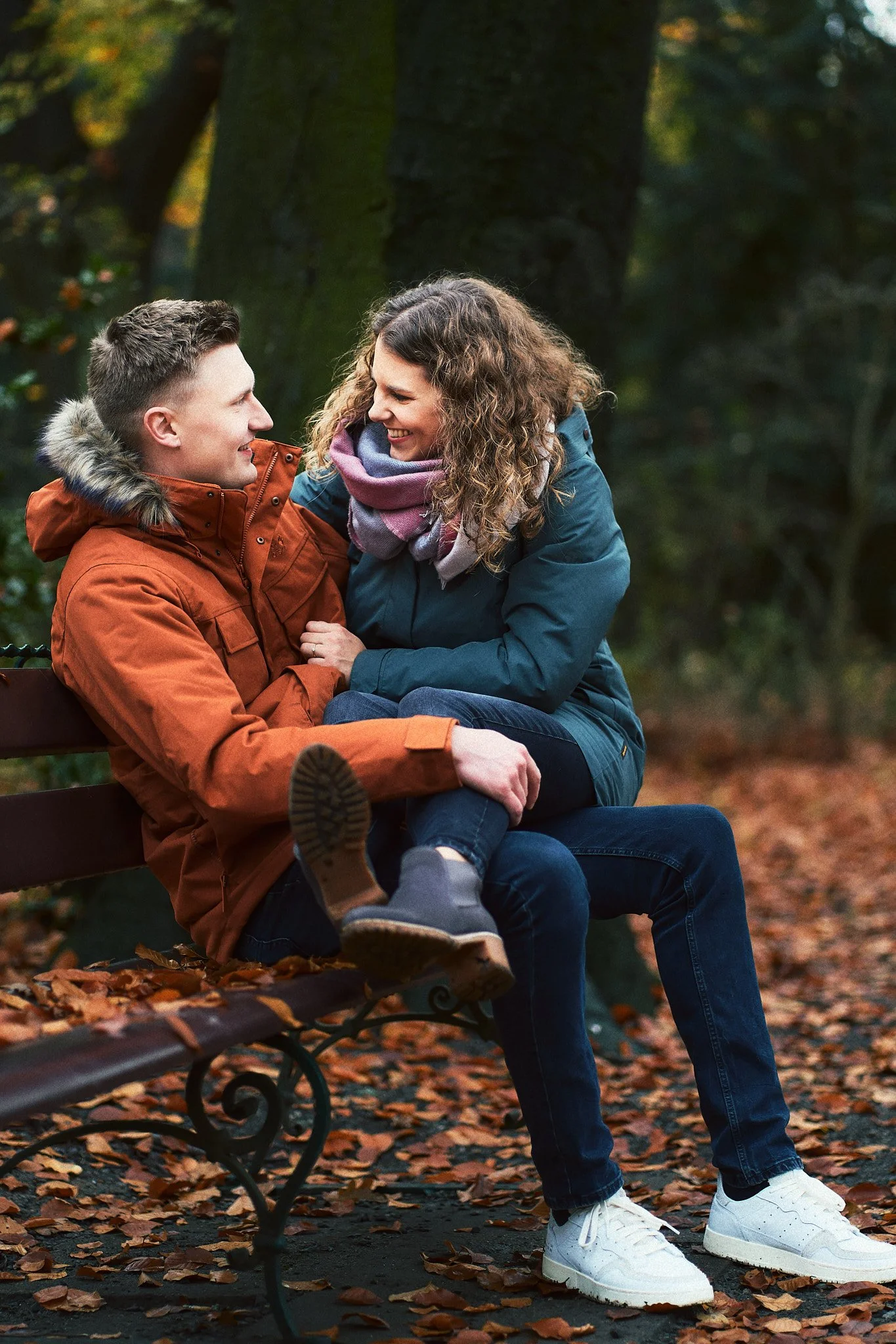 Ein lachendes Paar sitzt auf einer Parkbank, umgeben von Laub im Herbst, im Gespräch in einem grünen Park.