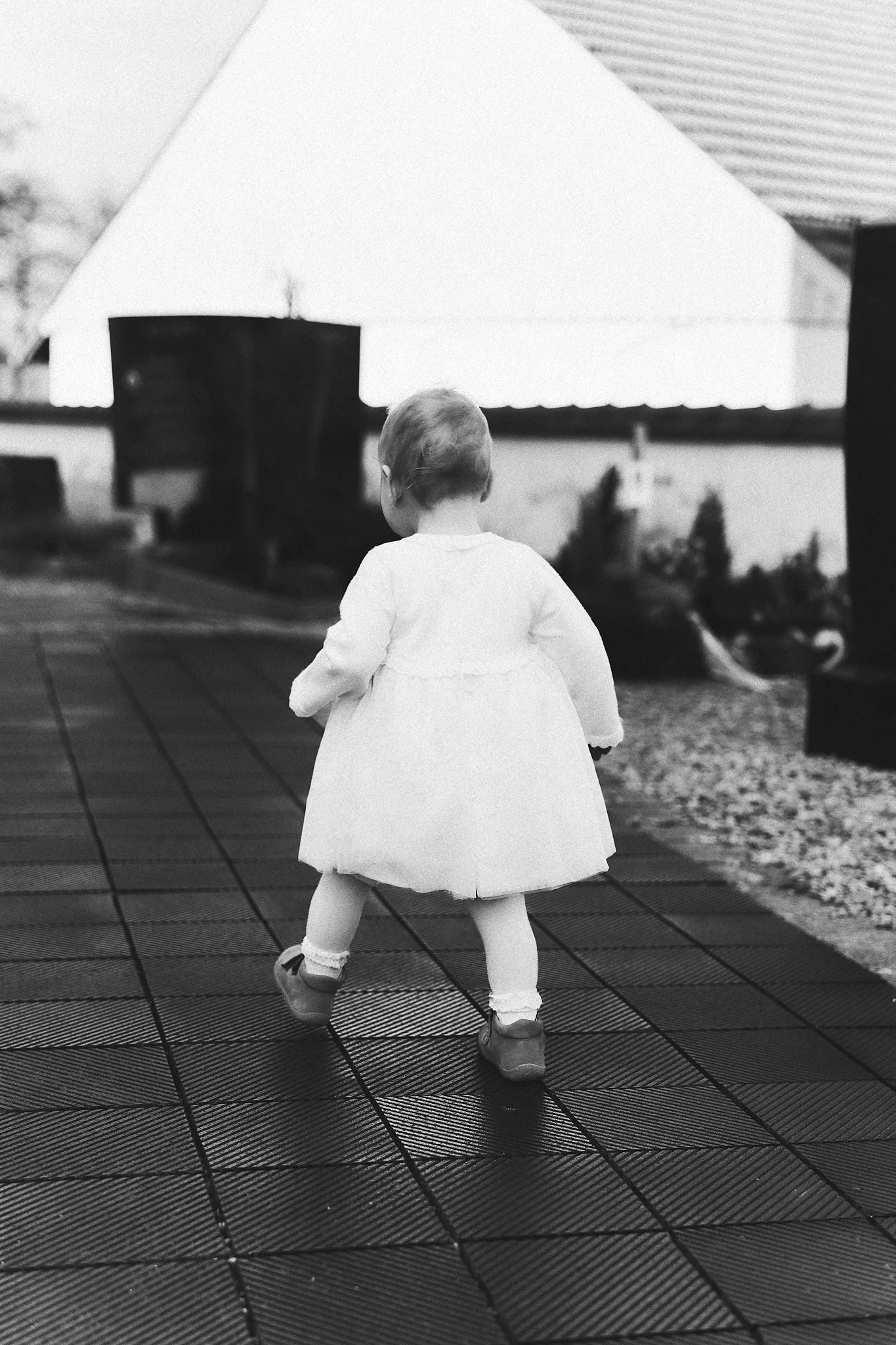 Ein kleines Mädchen mit kurzen Haaren, in einem weißen Kleid und Socken, läuft auf einer rutschfesten Oberfläche nach draußen.