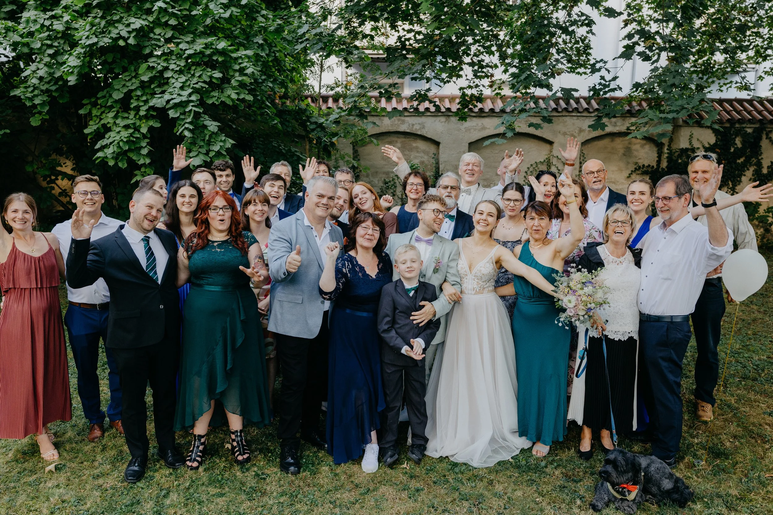Gruppe von Menschen bei einer Hochzeit im Garten, viele lachen und winken, ein Brautpaar in der Mitte, Blumen und ein Hund auf dem Gras