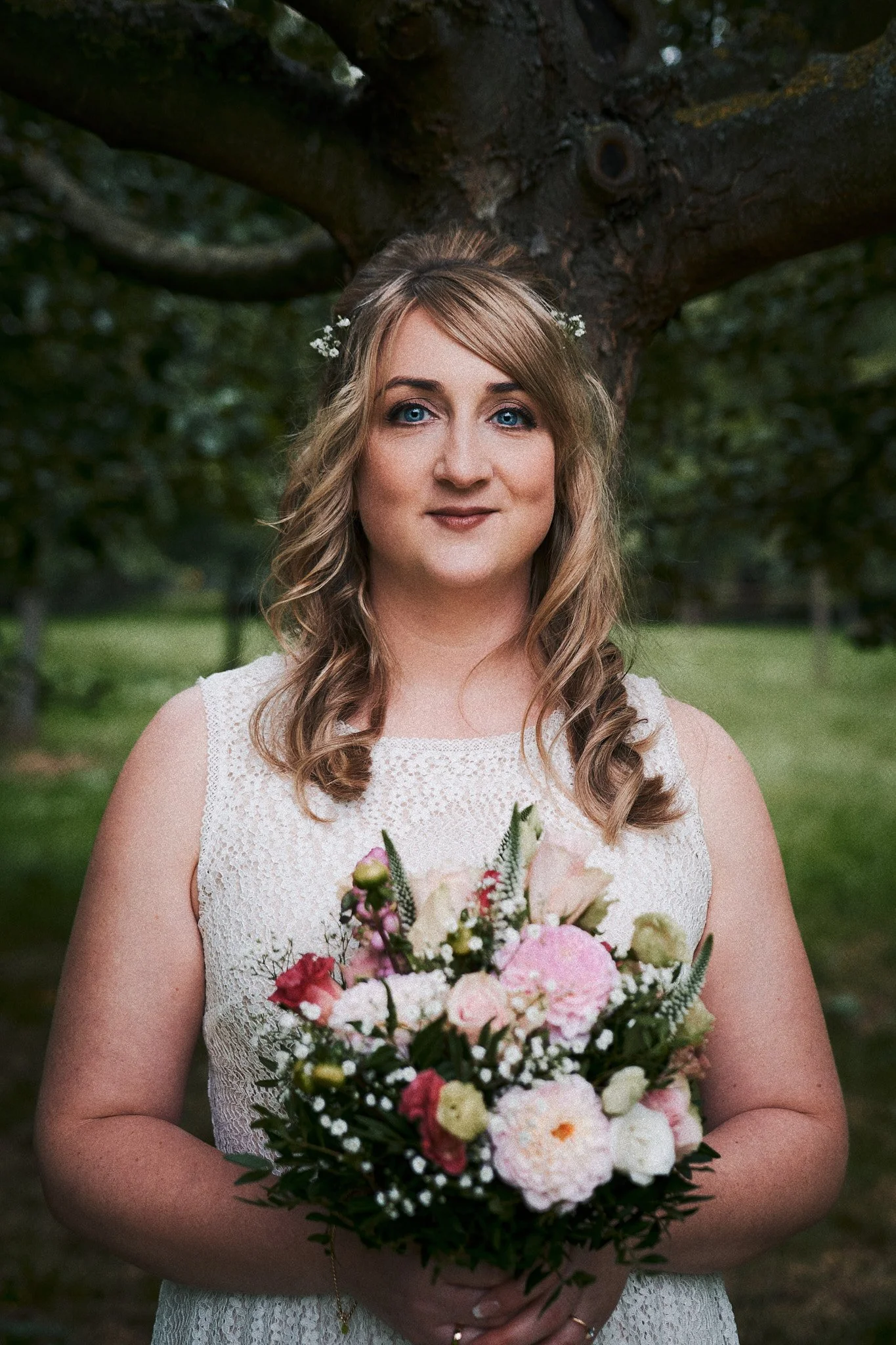 Eine Frau mit blonden, lockigen Haaren und blauen Augen hält einen bunten Blumenstrauß. Sie trägt ein weißes Spitzenkleid und befindet sich im Freien vor einem Baum mit grünen Blättern.