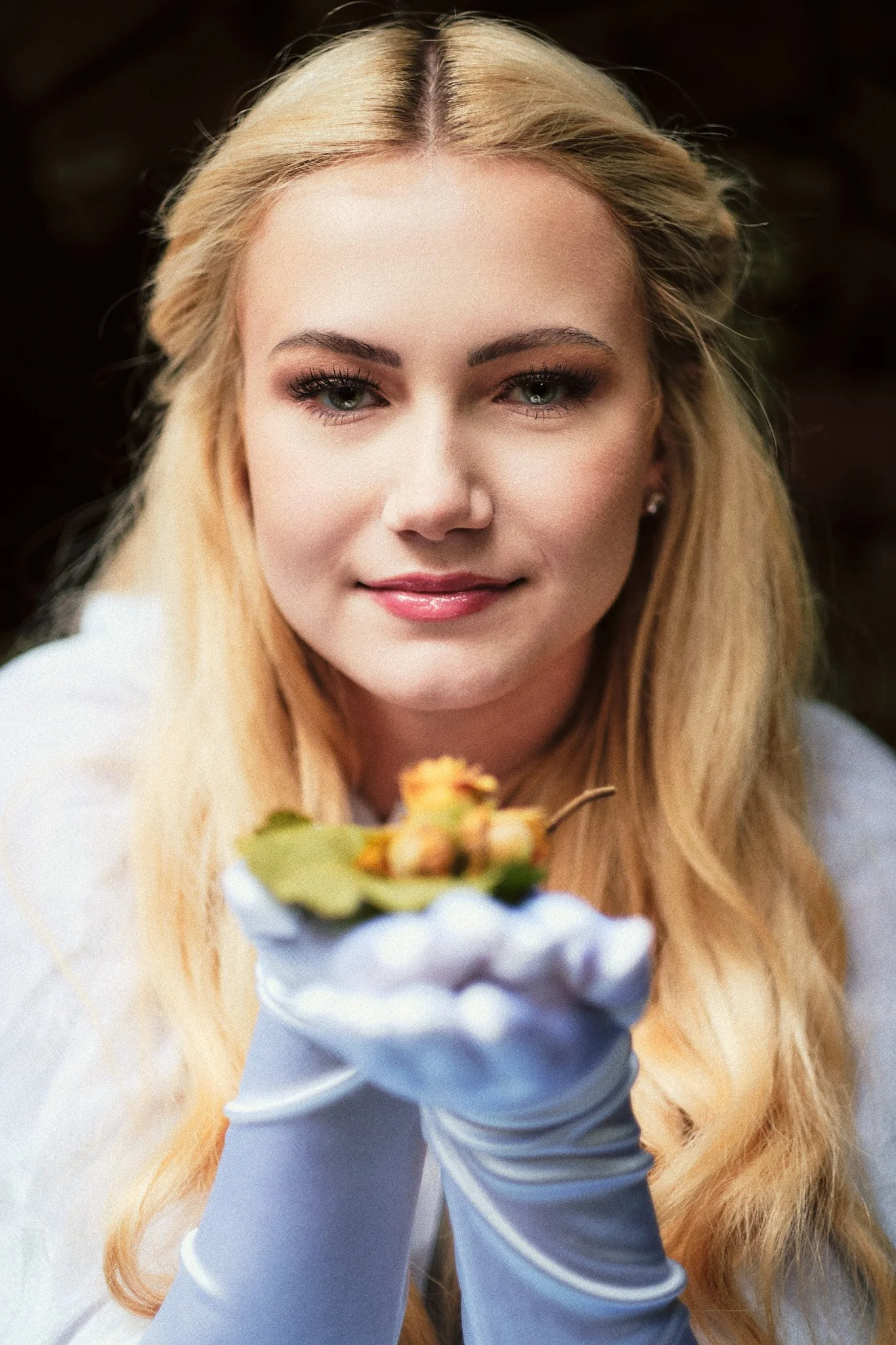 Junge Frau mit blonden Haaren, weißen Handschuhen, hält eine kleine Pflanze mit Blättern und Blüten, schaut freundlich in die Kamera.