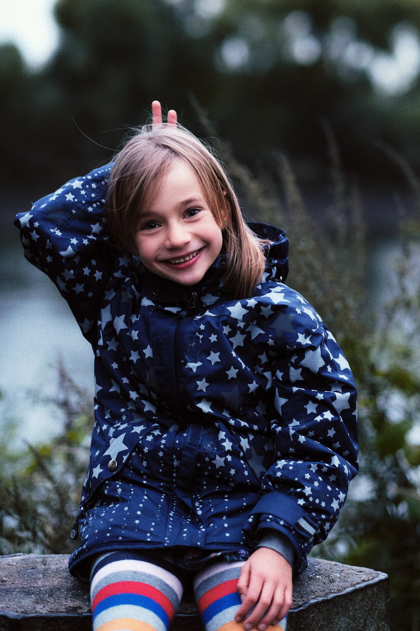 Ein lachendes Mädchen mit braunen Haaren, das auf einem Stein sitzt, trägt eine blaue Jacke mit weißen Sternen und bunte gestreifte Strümpfe, im Hintergrund Wald.