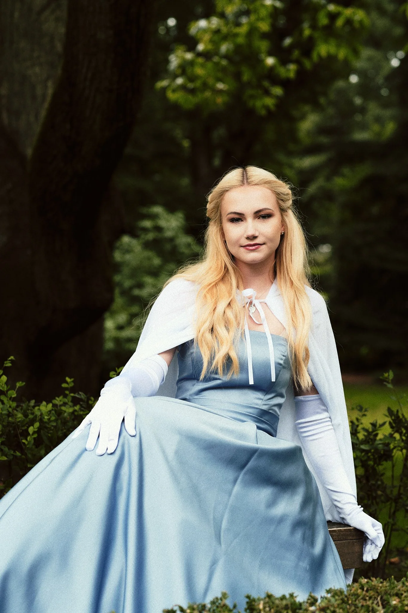Junge Frau in einer blauen Ballkleid und weißen Handschuhen sitzt auf einer Bank im Park, umgeben von grünen Büschen und Bäumen.
