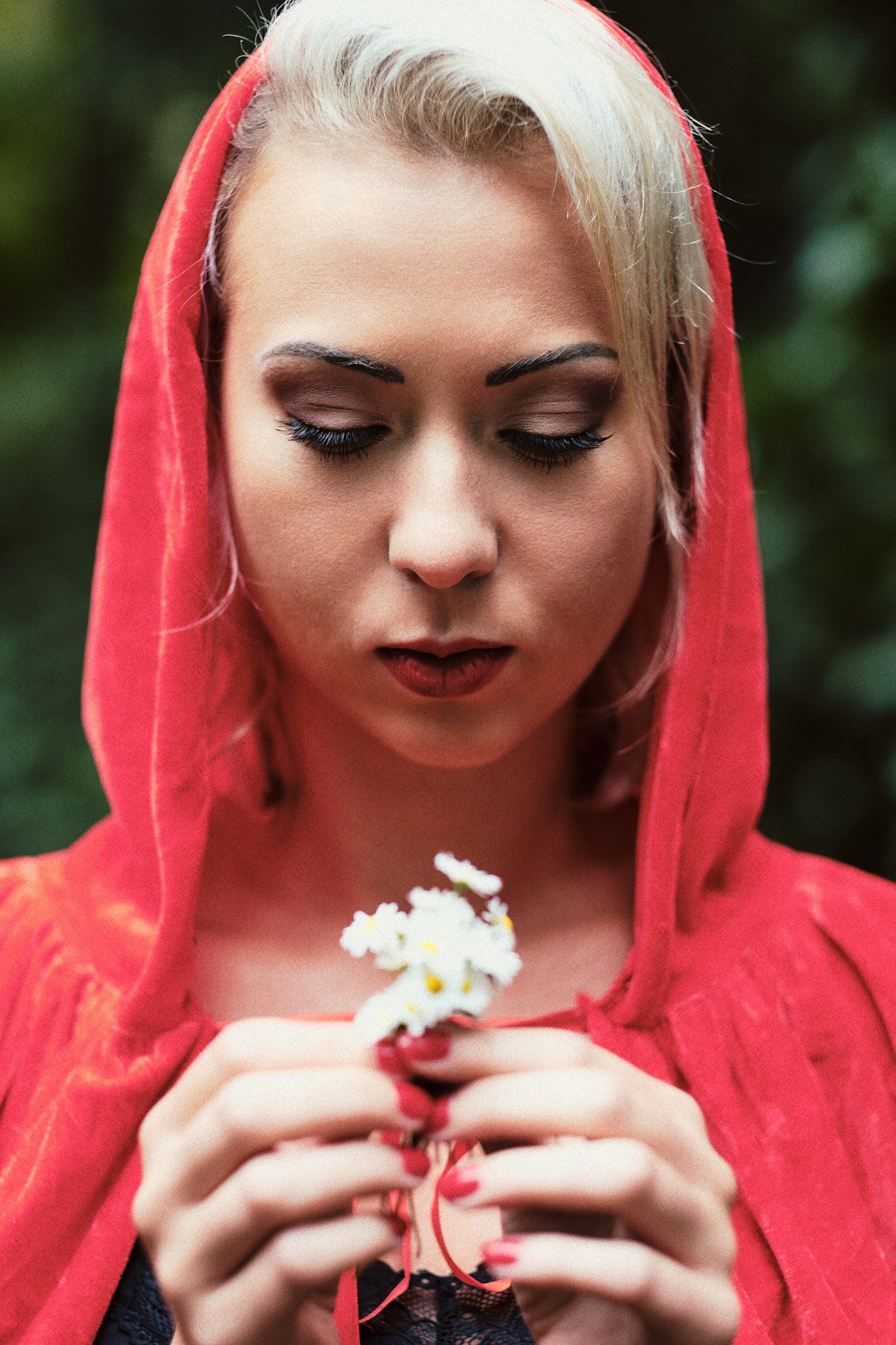 Junge Frau mit blondem Haar, rotem Kapuzenumhang, hält kleine weiße Blumen mit gelben Zentren in den Händen, im Hintergrund unscharfes grünes Laub.