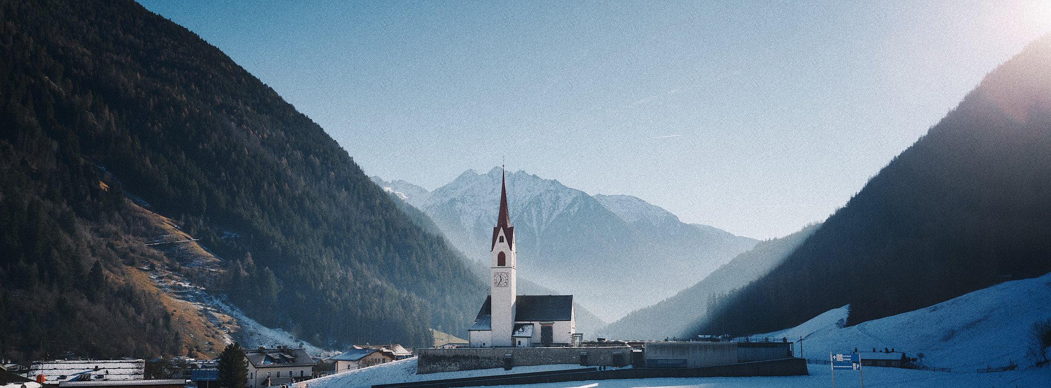 Eine verschneite Kirche in einem Tal, umgeben von hohen, bewaldeten Bergen bei Sonnenaufgang oder -untergang in den Alpen.