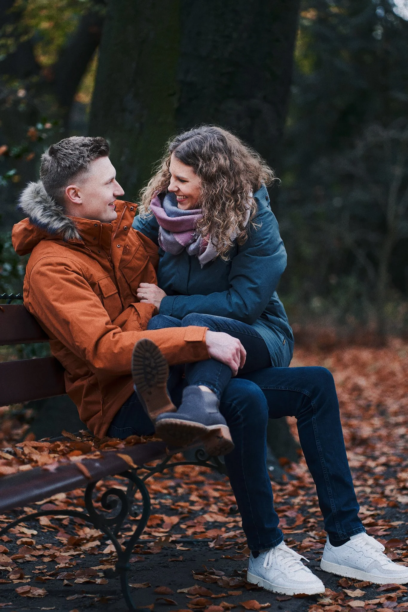 Zwei Menschen sitzen auf einer Parkbank im Herbst, lachen und schauen sich liebevoll an, während Blätter auf dem Boden liegen.