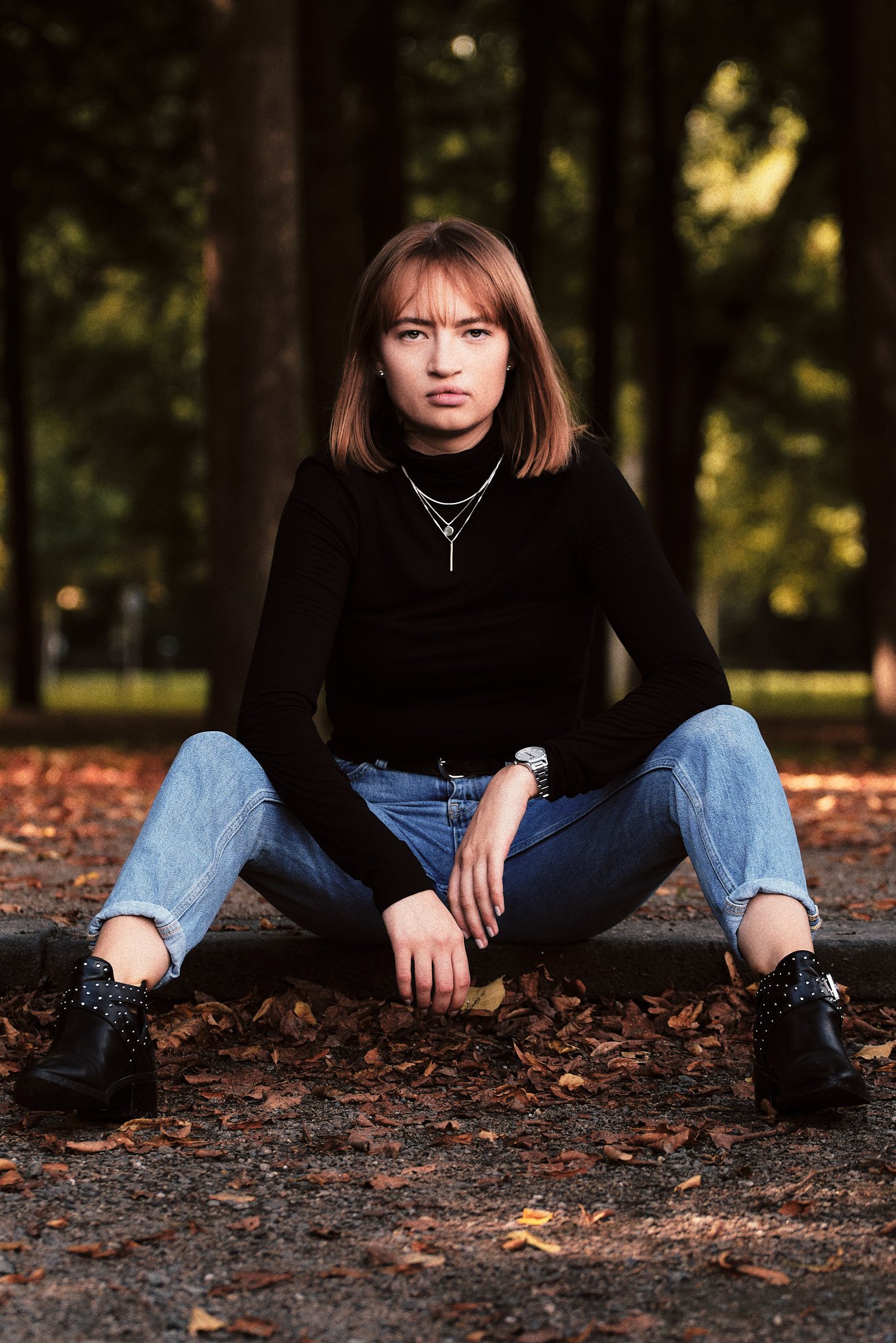 Junge Frau sitzt auf dem Boden in einem Park, umgeben von Bäumen, trägt schwarze Stiefeletten, blaue Jeans und schwarzen Pullover, mit Schmuck und Uhren, Blick in die Kamera gerichtet.