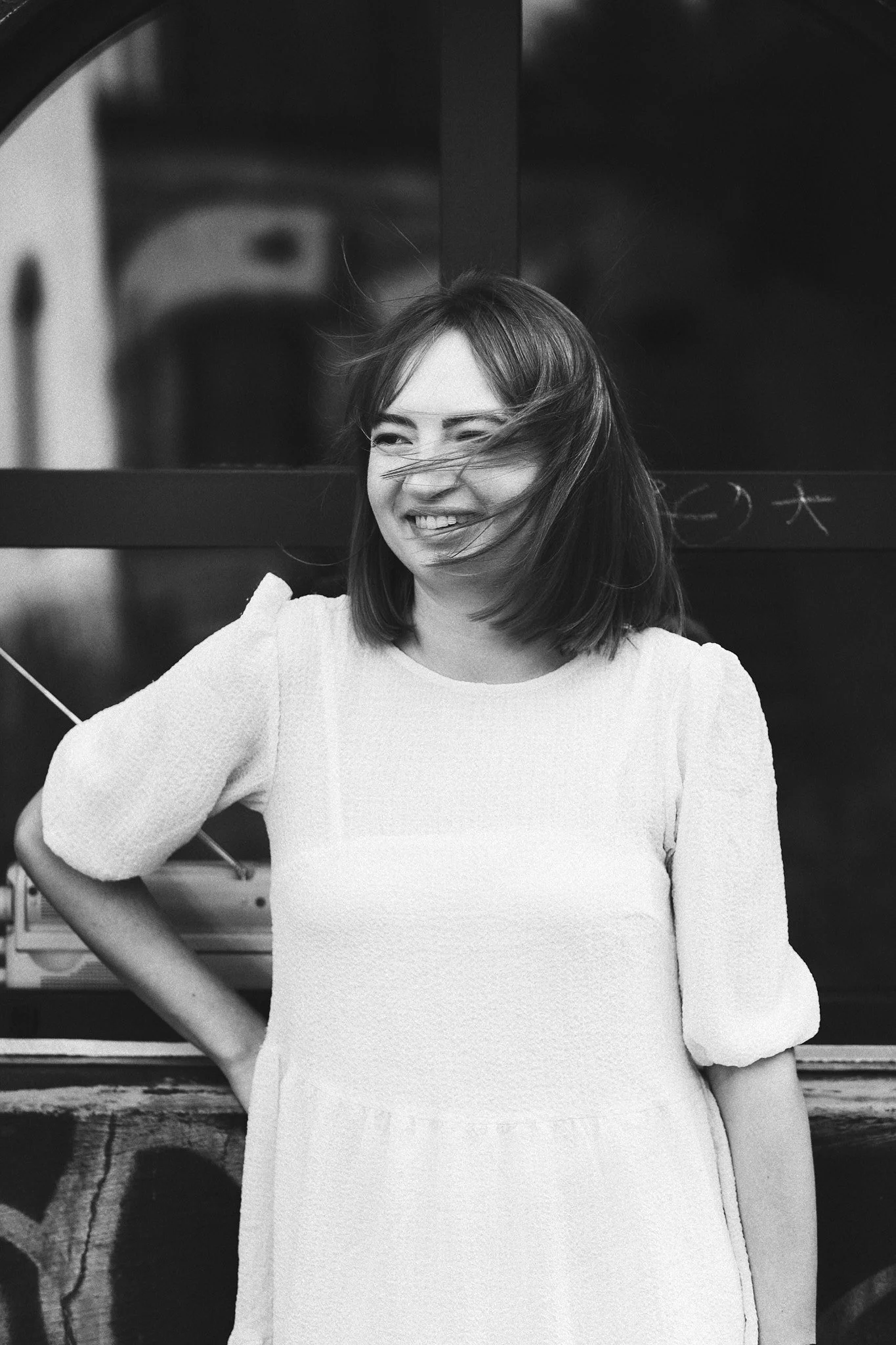 Junge Frau lacht und lächelt bei Wind, gestikuliert mit der Hand, trägt ein weißes Kleid, steht vor einem dunklen Hintergrund.