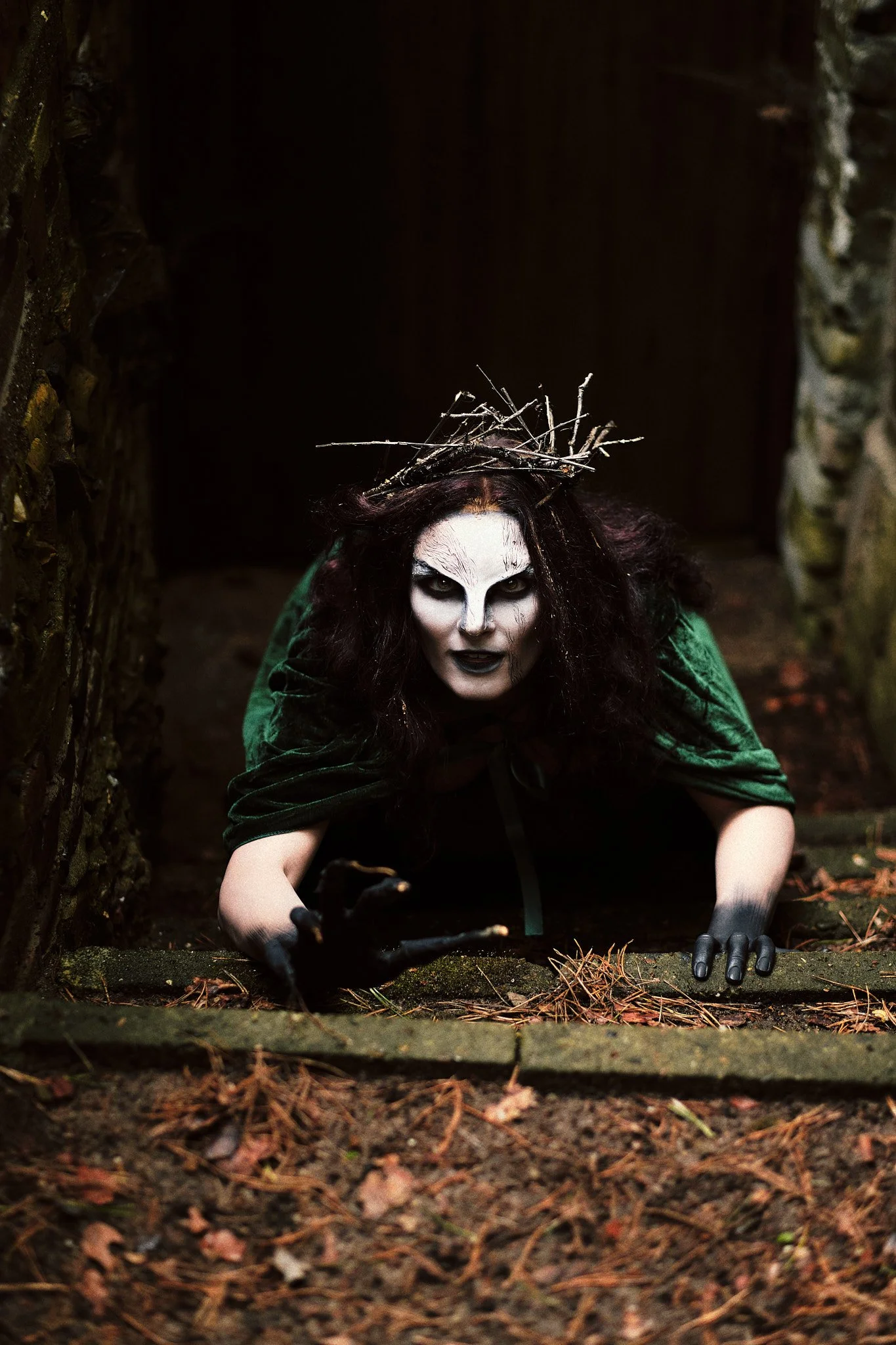 Eine gruselige Figur mit weißem Gesicht, schwarzen Augen und dunklem Haar, gekleidet in grünem Umhang und schwarzen Handschuhen, kriecht aus einem dunklen, alten Durchgang im Wald.