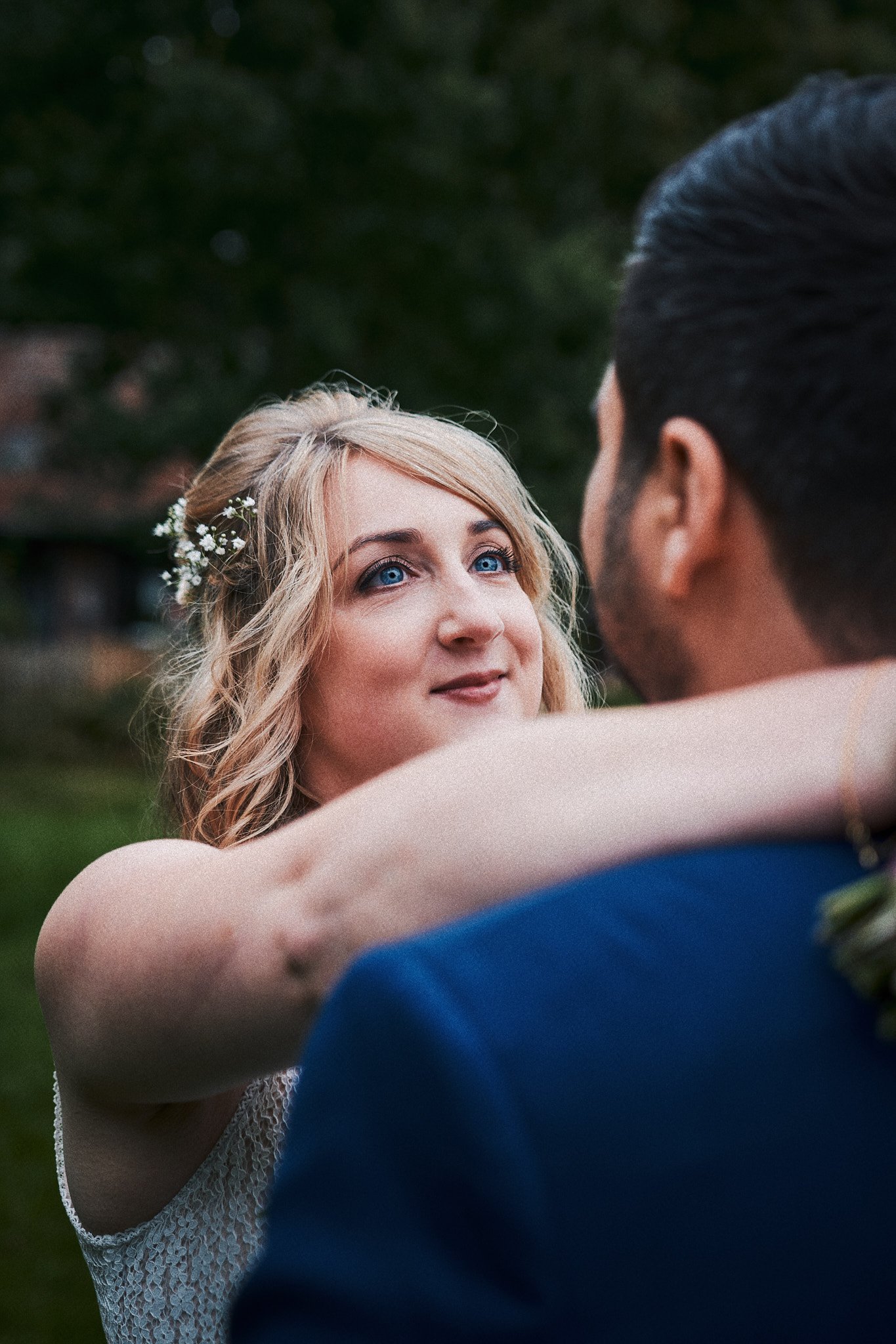 Ein blondes Mädchen mit blauen Augen und weißen Blumen im Haar schaut in die Augen eines Mannes, während sie sich umarmen, im Freien bei Dämmerlicht.