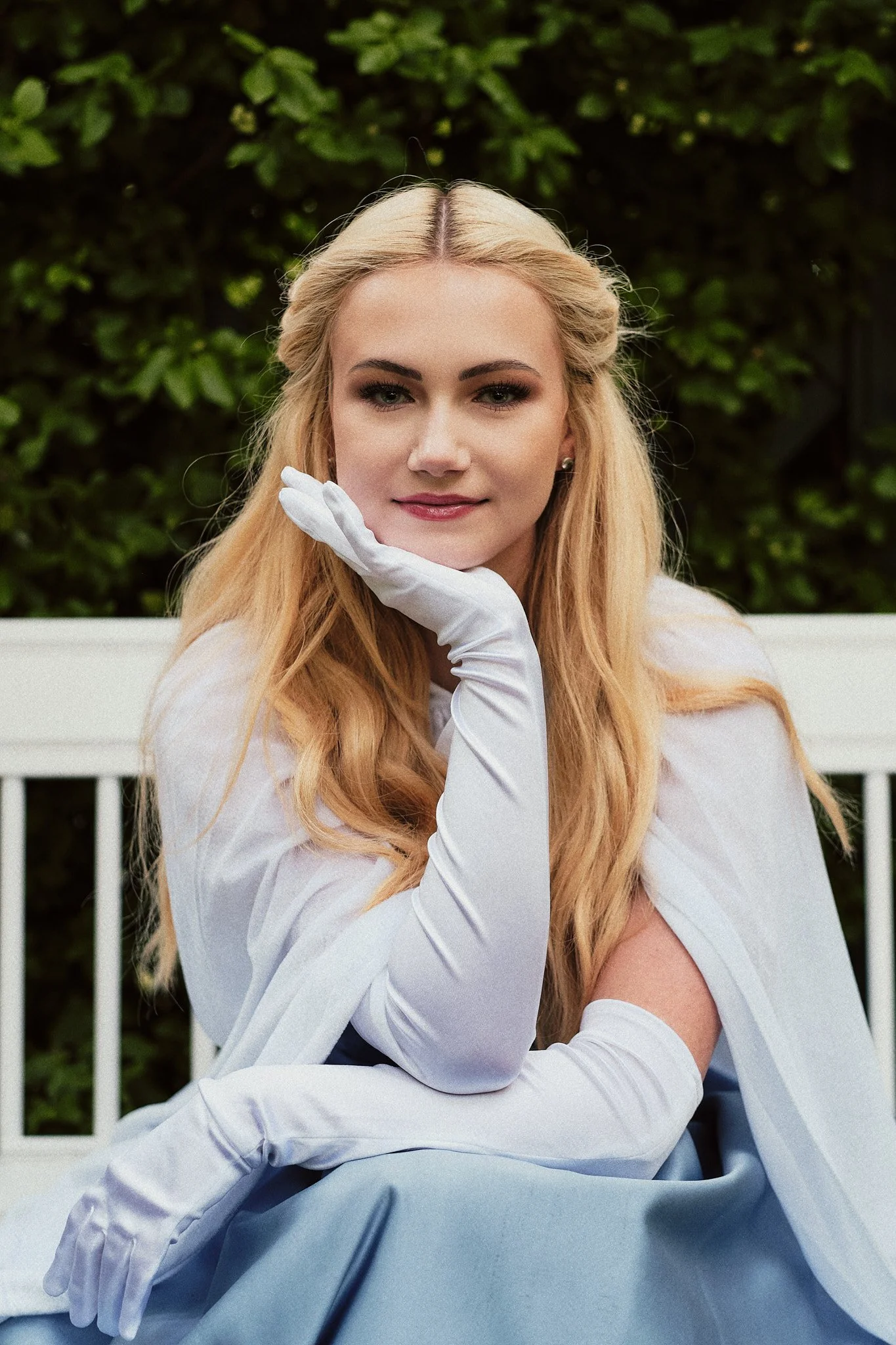 Eine junge Frau mit langen, blonden Haaren trägt elegante blaue und weiße Kleidung und weiße Handschuhe. Sie sitzt auf einer weißen Bank im Freien vor einer grünen Gartenterrasse, posiert mit ihrem Kopf auf ihrer Hand und lächelt leicht in die Kamera