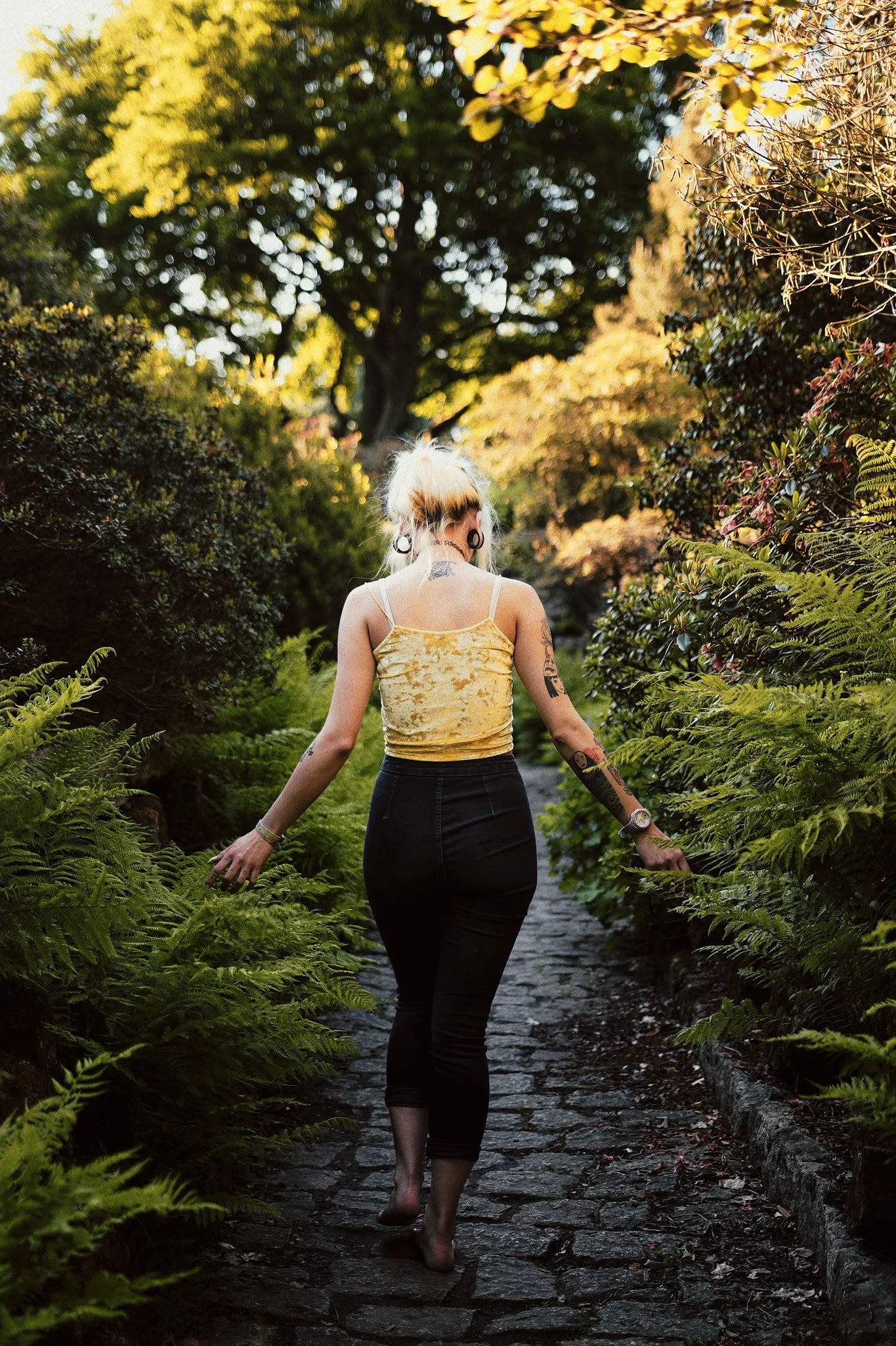 Eine junge Frau mit Tattoos, blondem Haar in einem hohen Pferdeschwanz, barfuß auf einem steinigen Weg in einem Garten mit grünen Pflanzen und Bäumen, bei Sonnenuntergang.