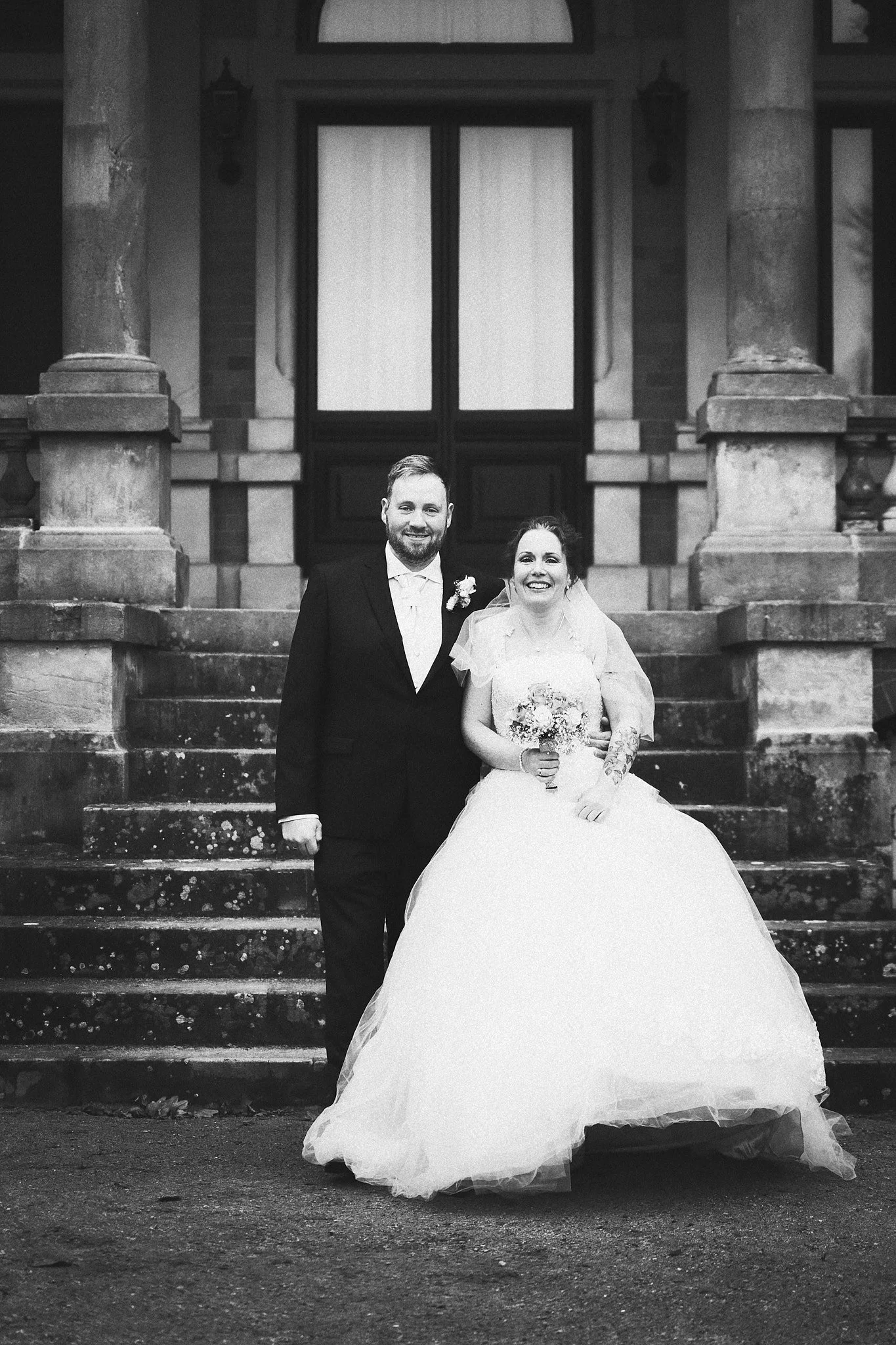Ein Hochzeitsfoto eines Paares, das vor einer alten Stein-Treppe und einem historischen Gebäude steht. Der Mann trägt einen schwarzen Anzug mit weißen Hemd und Fliege, die Frau trägt ein weißes Hochzeitskleid und hält einen Blumenstrauß.