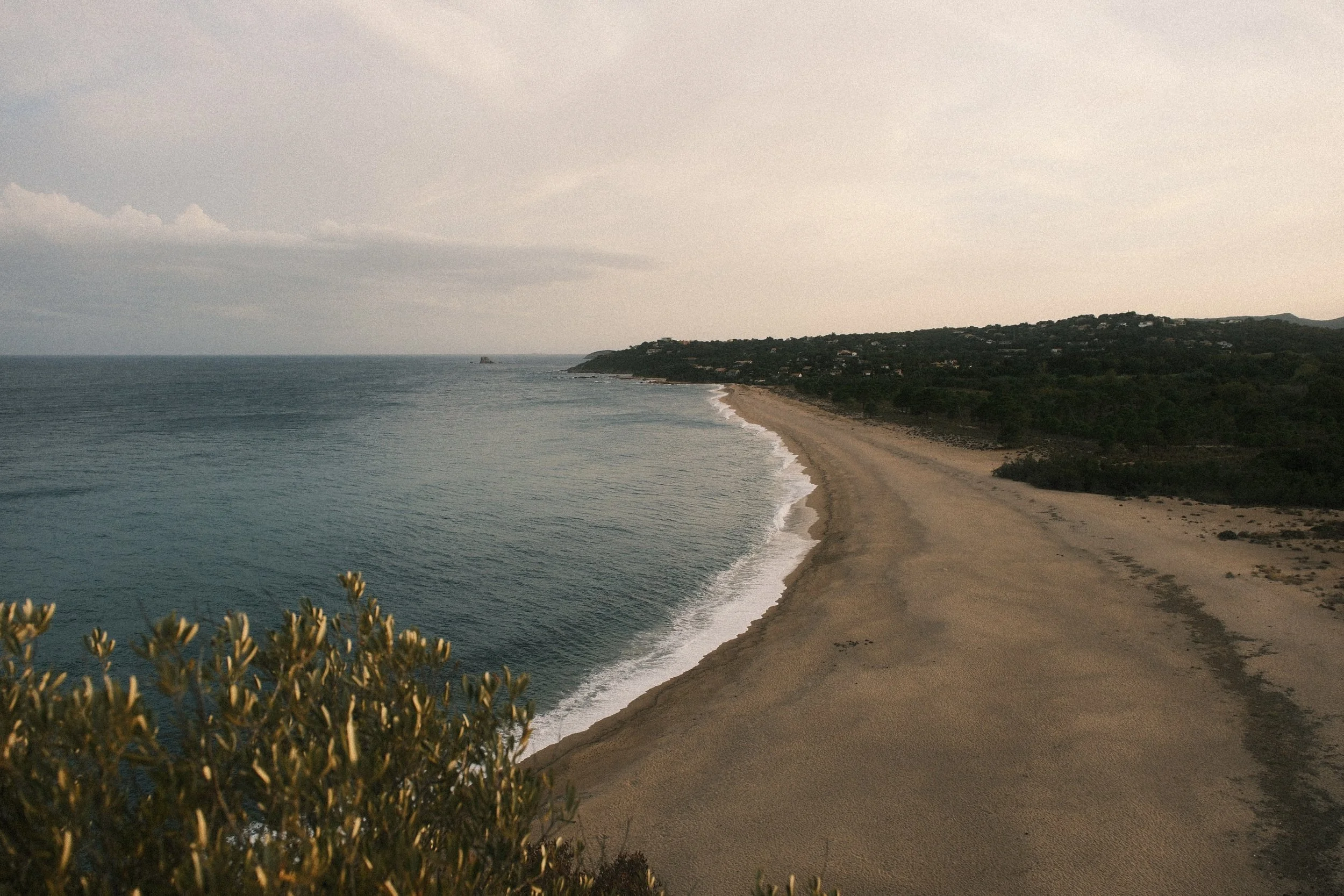 Ein Strand bei Sonnenuntergang mit Sand, Wasser und einem bewaldeten Hügel im Hintergrund.