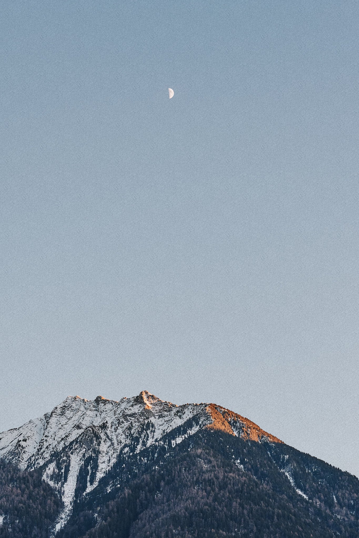 Berg mit schneebedecktem Gipfel und dunklem bewaldetem Hang unter blauem Himmel mit Halbmond