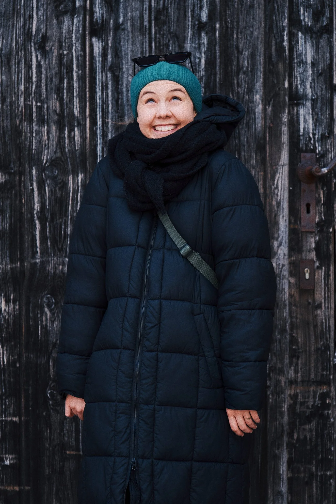 Junge Frau trägt eine blaue Mütze, eine schwarze Jacke und einen schwarzen Schal, steht vor einem Holzzaun, lächelt und schaut nach oben.