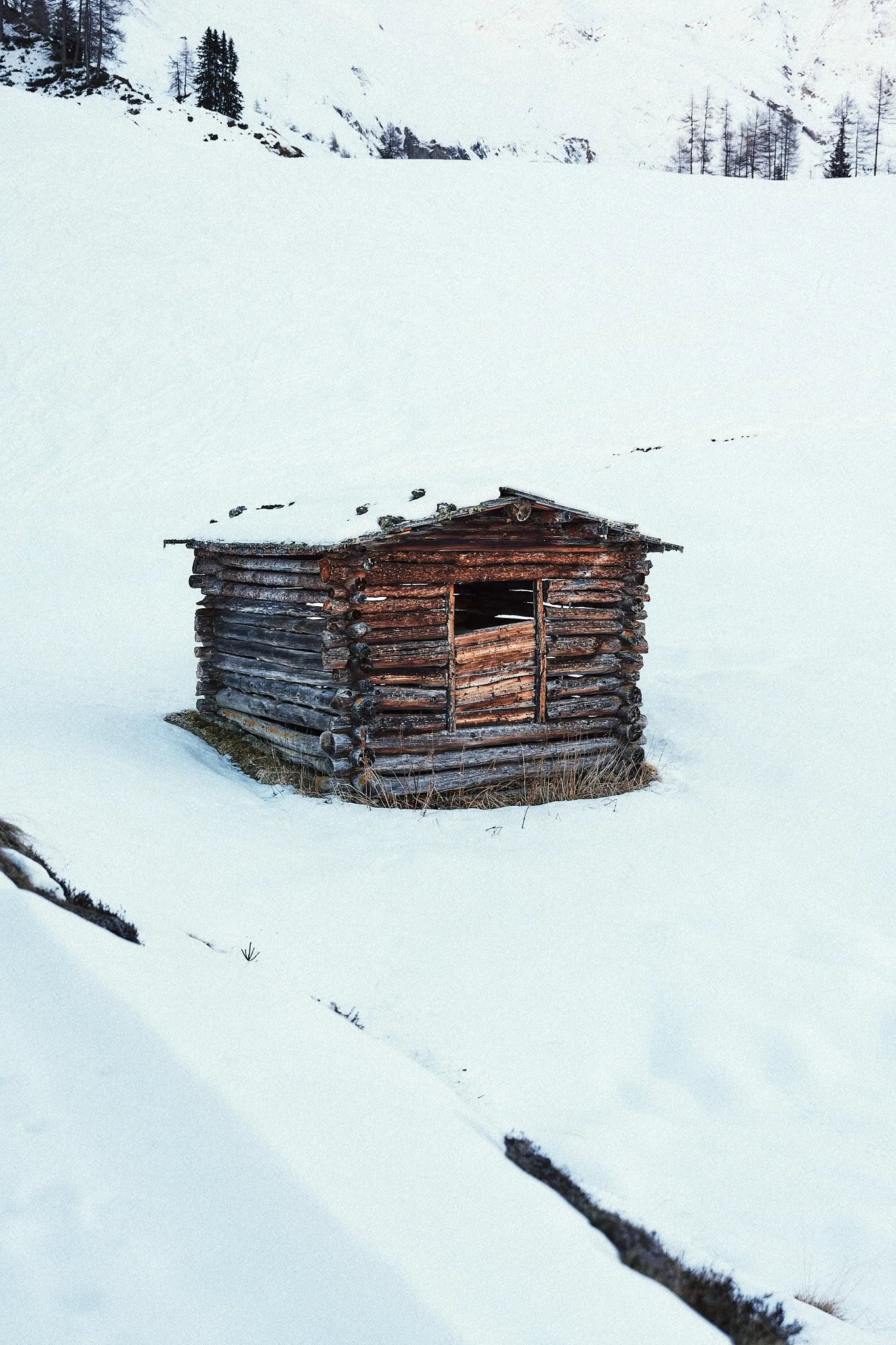 Ein kleines, altes aus Holz gebautes Haus in einer verschneiten Landschaft, umgeben von schneebedeckter Wiese und Bergen im Hintergrund.
