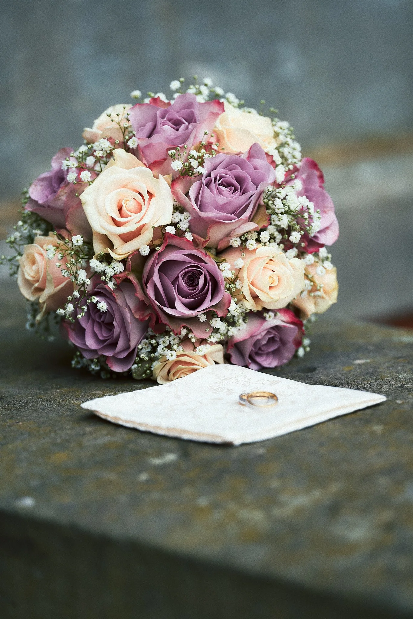 Ein Brautstrauß mit rosa und lila Rosen, kleinen weißen Blumen, liegt auf einem grauen Stein. Daneben liegt ein weißes Taschentuch mit zwei Eheringen.