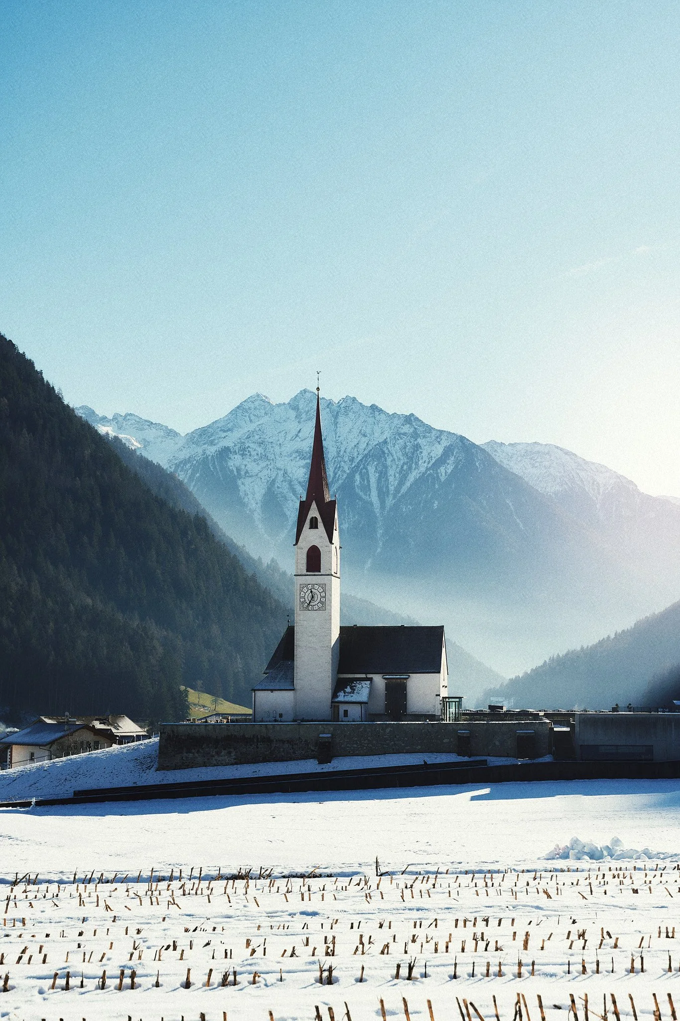 Eine weiße Kirche mit hoorem roten Turm vor schneebedeckten Feldern und in den Bergen im Hintergrund.