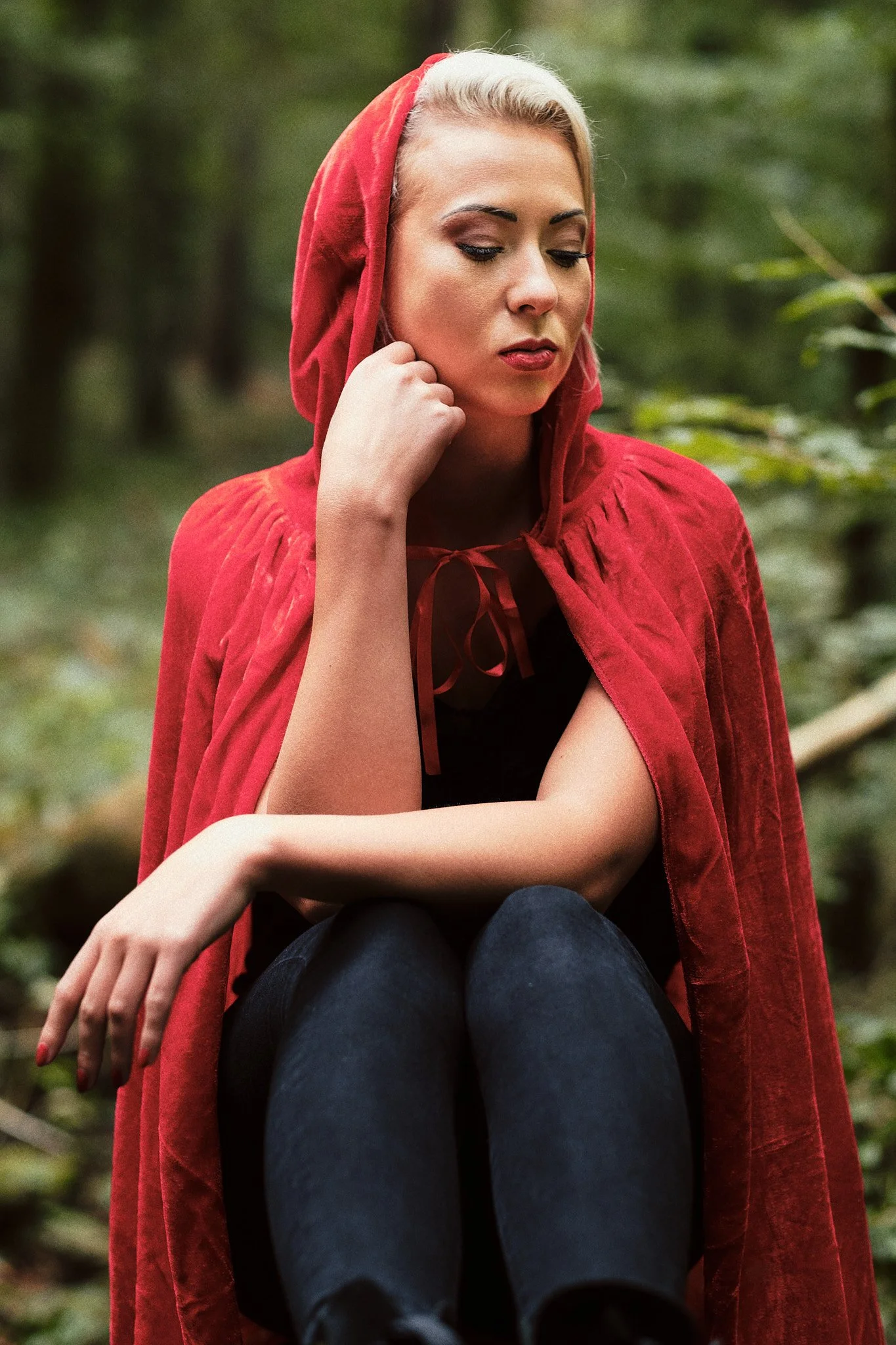 Junge Frau mit blondem Haar in einem roten Umhang sitzt im Wald und blickt nach unten.