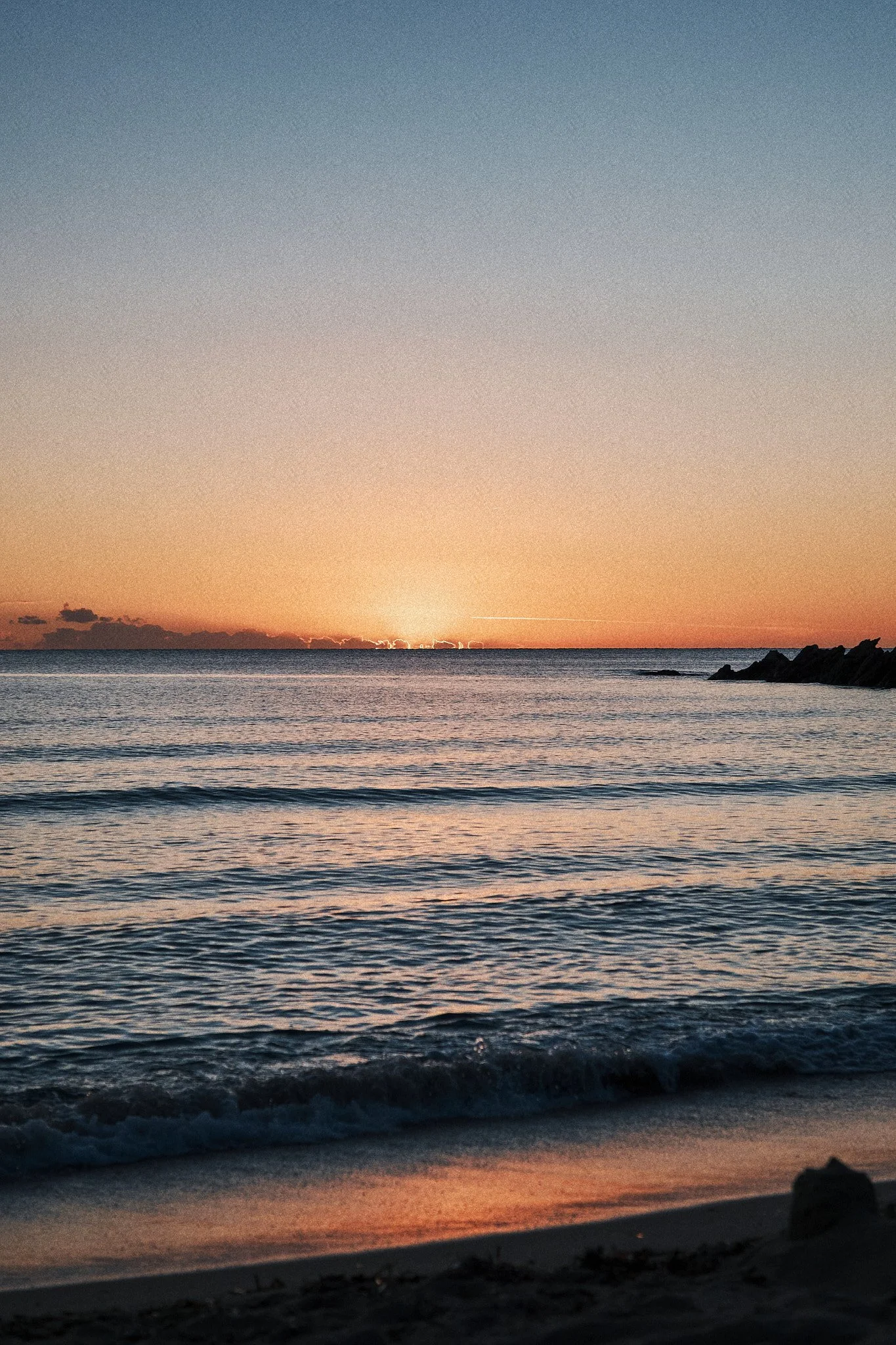 Sonnenuntergang am Meer mit ruhigen Wellen und Felsen in der Ferne.