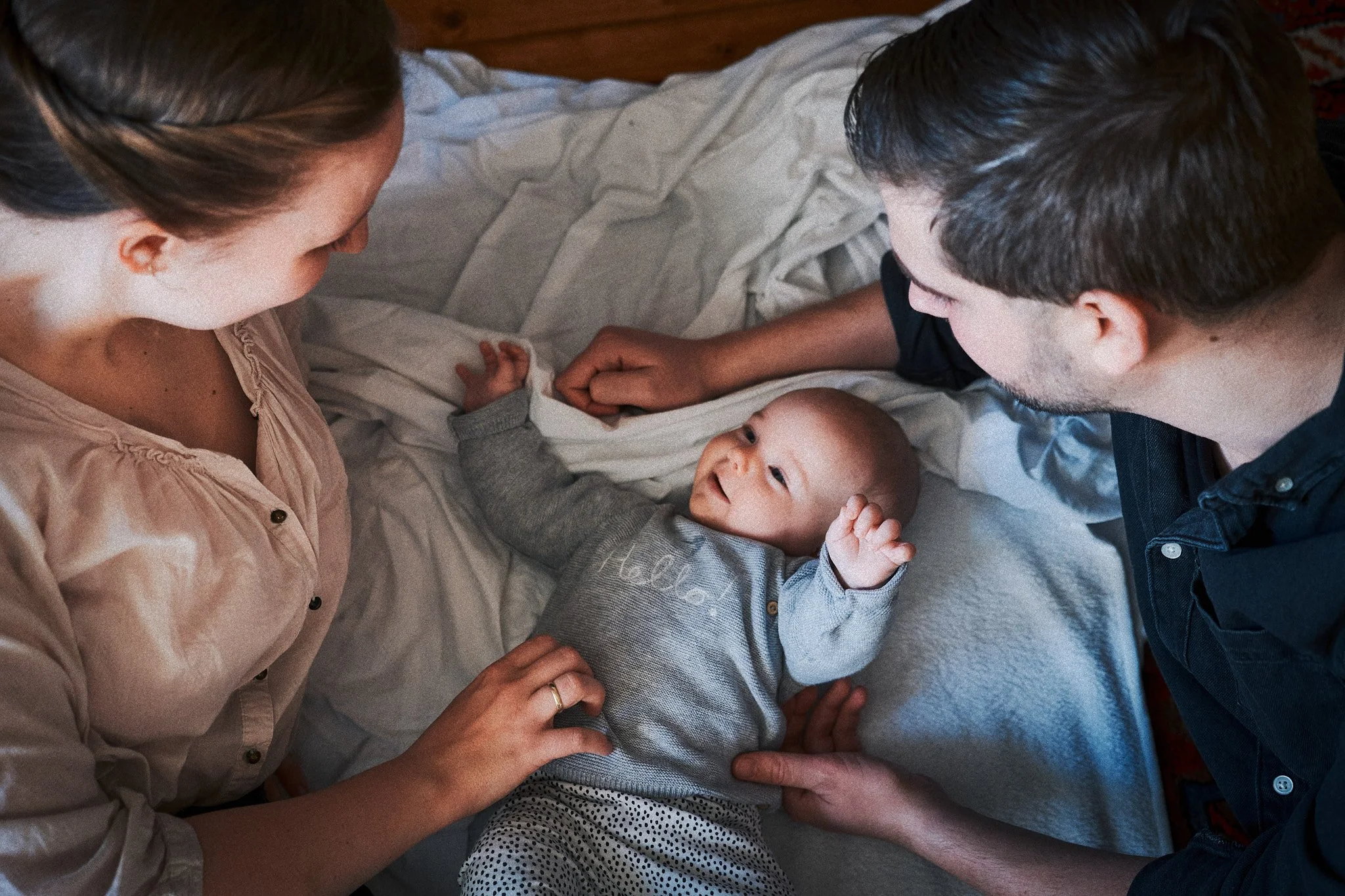 Ein Baby liegt auf einem Bett zwischen einem Mann und einer Frau. Das Baby schaut glücklich zu den Erwachsenen, die es liebevoll ans schauen.