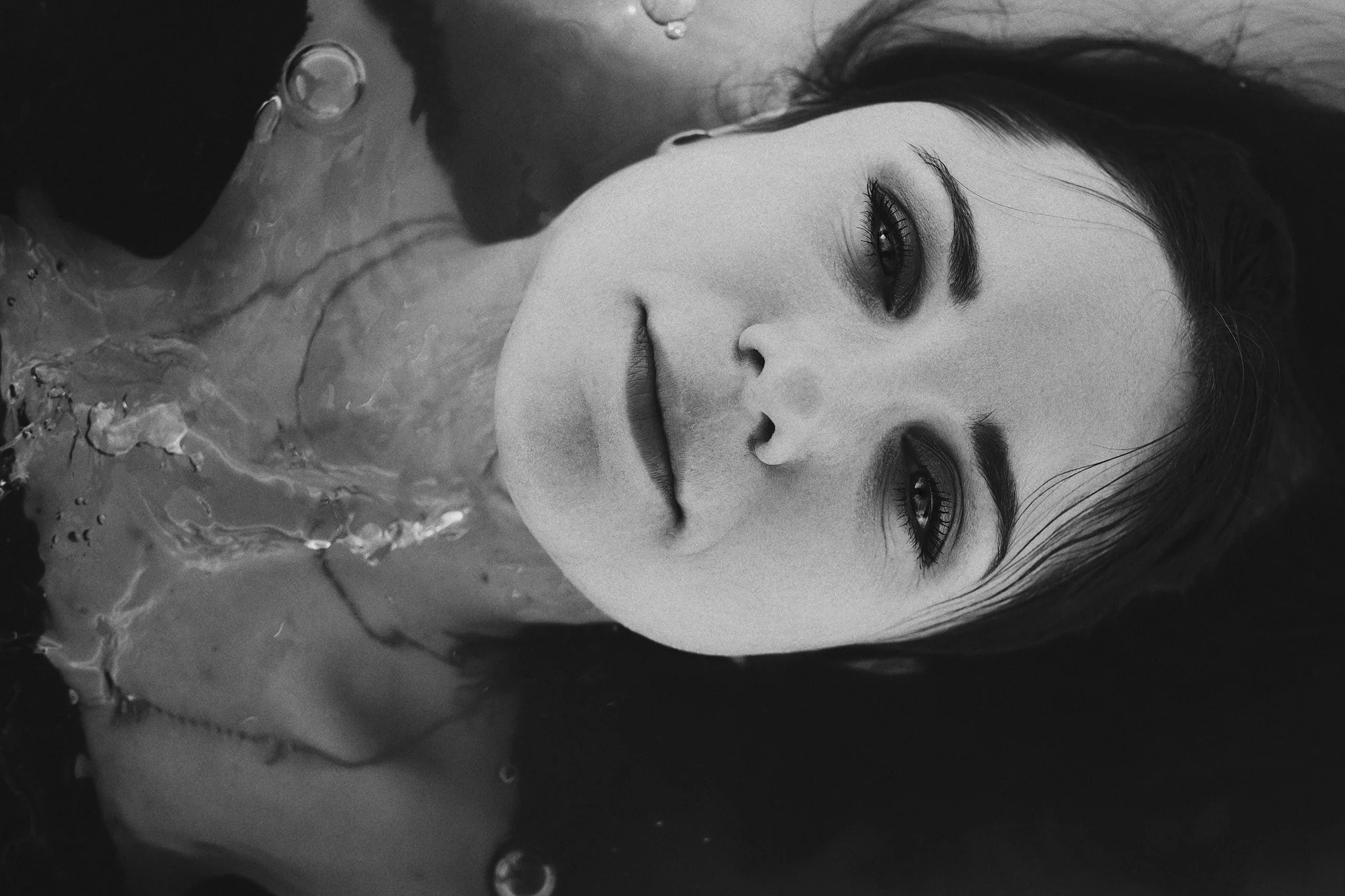 Schwarze und weiße Nahaufnahme eines liegenden Frauengesichts mit geöffneten Augen und nassem Haar, umgeben von Wasser.