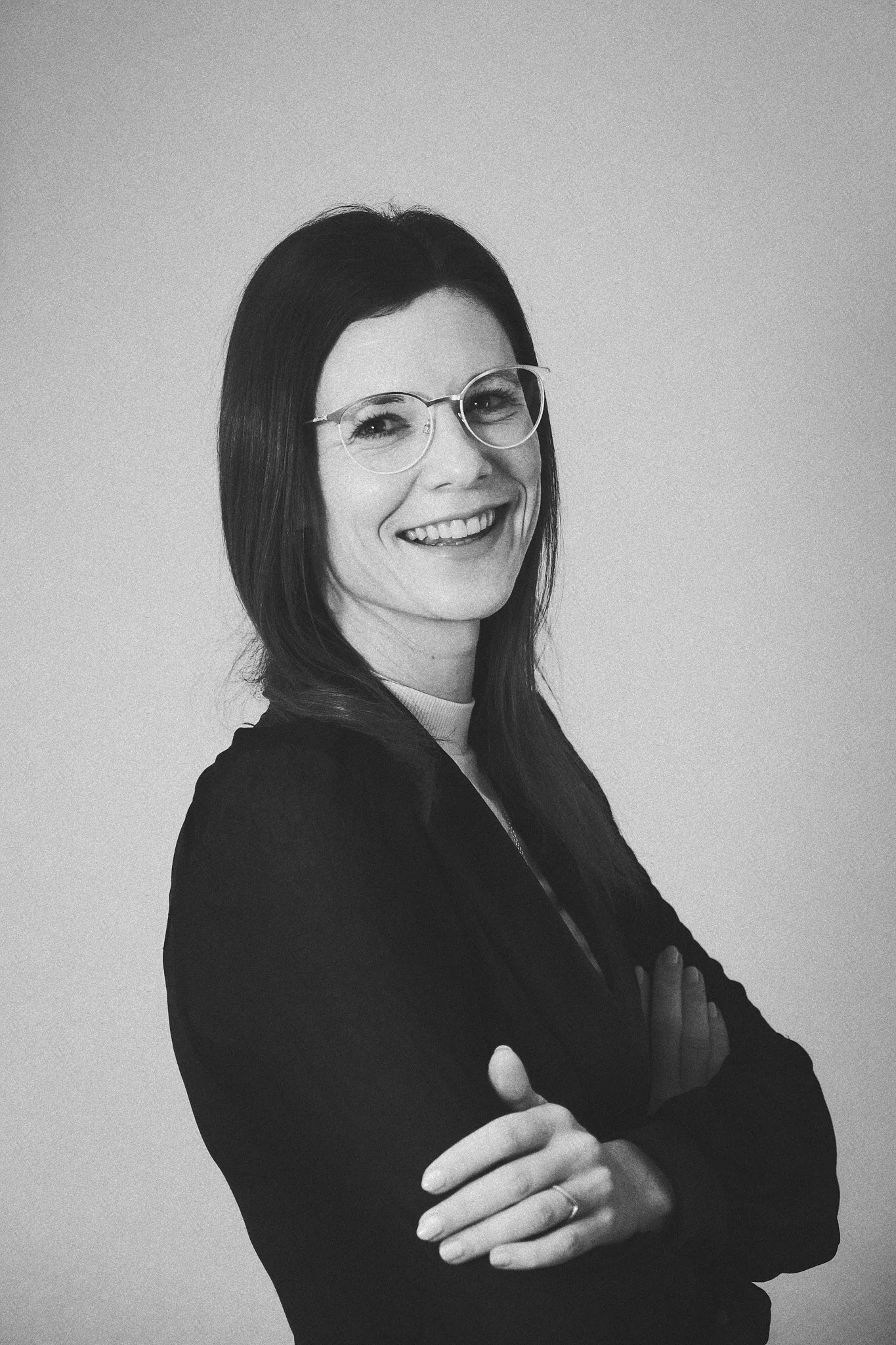 Schwarz-weiß Portrait einer lachenden Frau mit schwarzen Haaren und Brille, die Arme verschränkt.